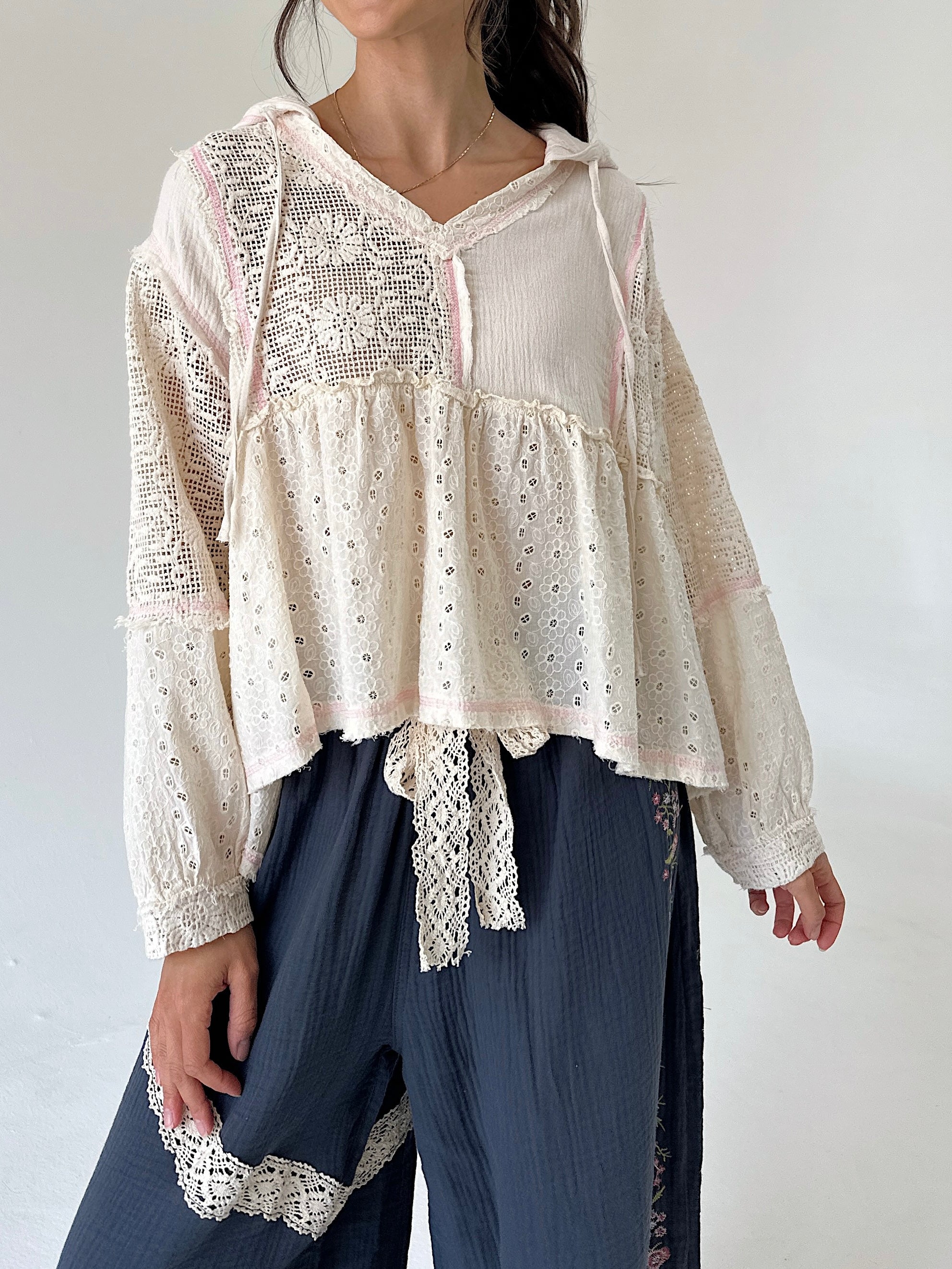 A woman wearing a cream eyelet lace long-sleeve blouse with a tie waist and dark blue wide-leg pants against a plain white background.