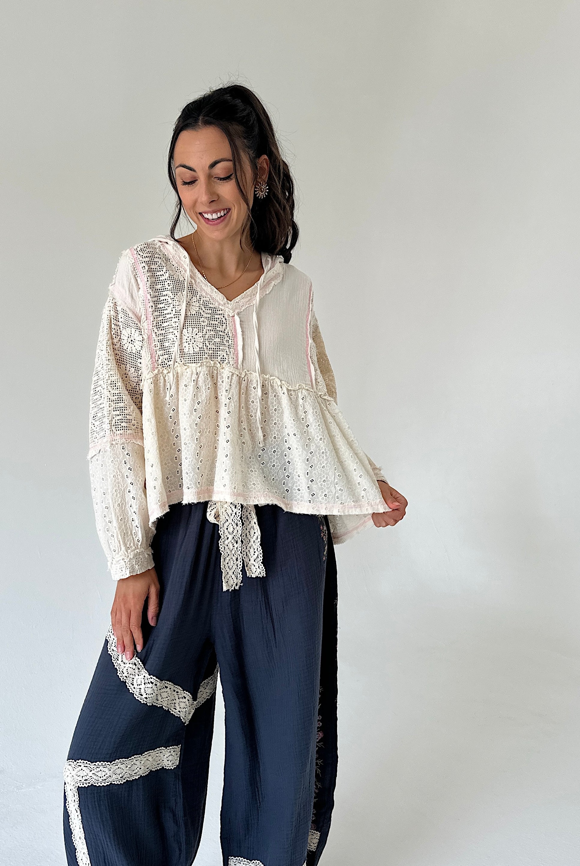 A woman modeling a cream-colored bohemian-style blouse with intricate eyelet embroidery and a ruffled peplum hem, paired with navy blue wide-leg pants featuring lace trim, set against a plain light gray studio background.