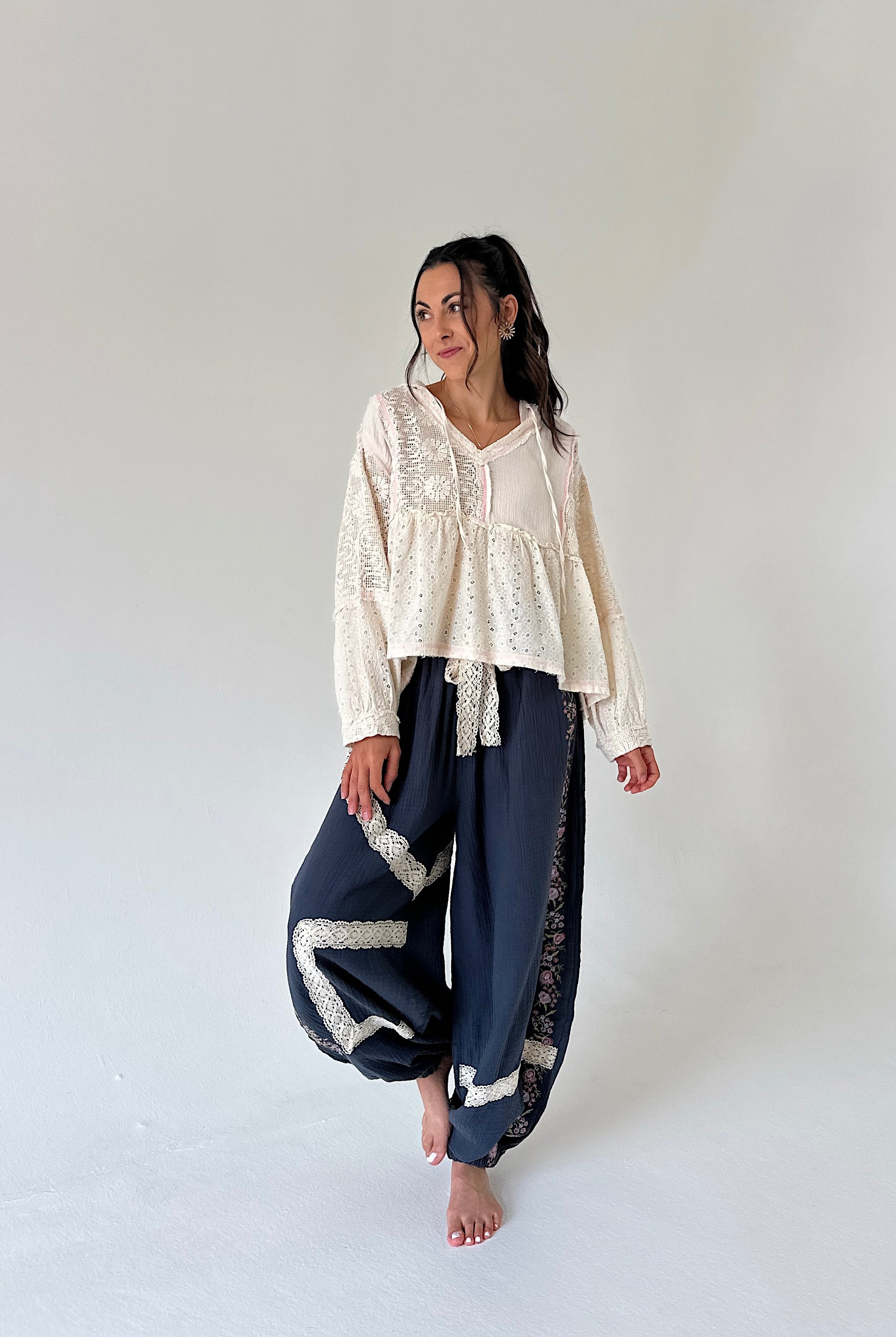 A woman stands barefoot in a studio wearing a cream lace long-sleeve top and navy blue wide-leg pants with white lace trim.