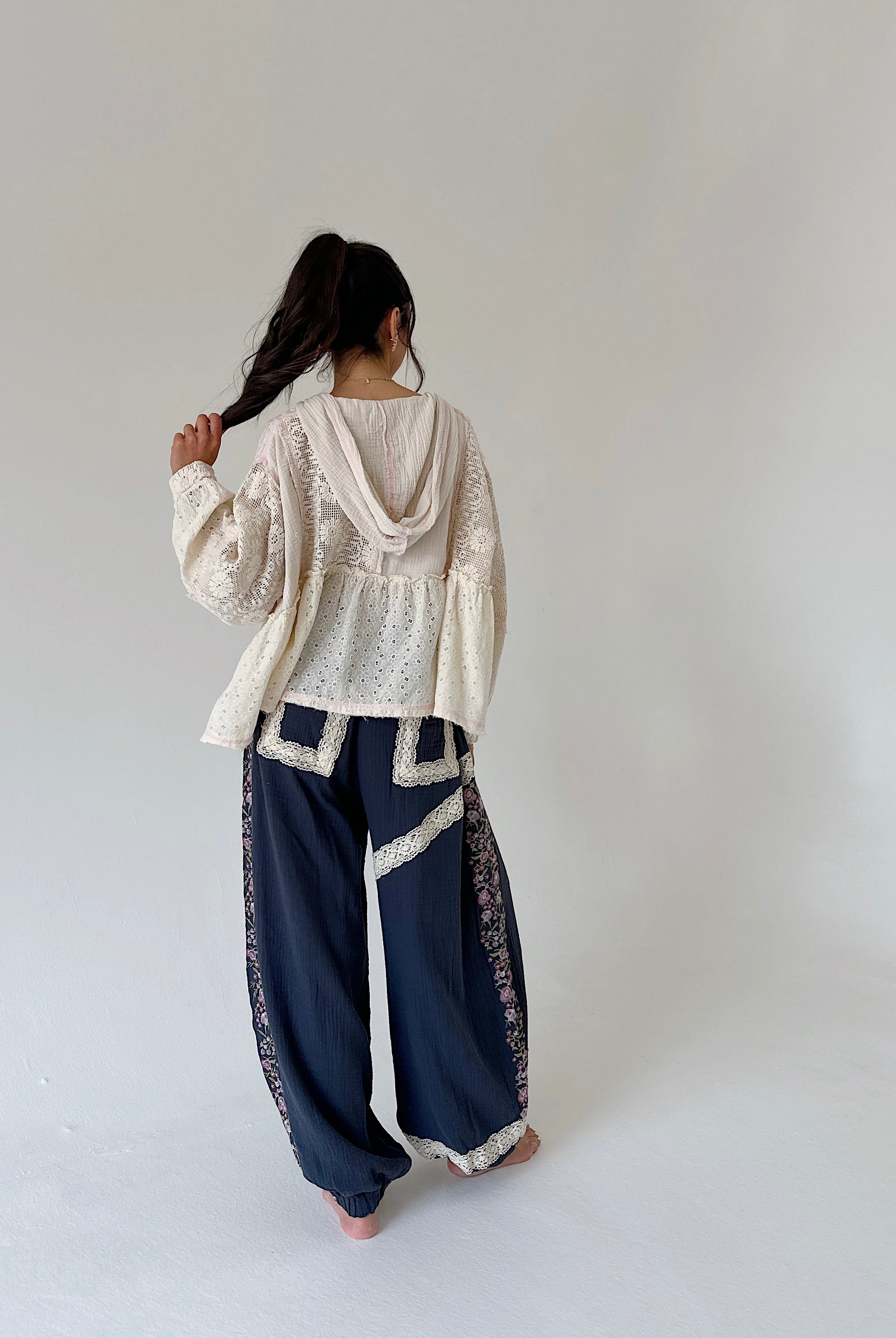A woman stands barefoot against a plain light gray studio background, viewed from behind. She is wearing a cream lace long-sleeve top and loose dark navy wide-leg pants with cream trim and side floral embroidery.