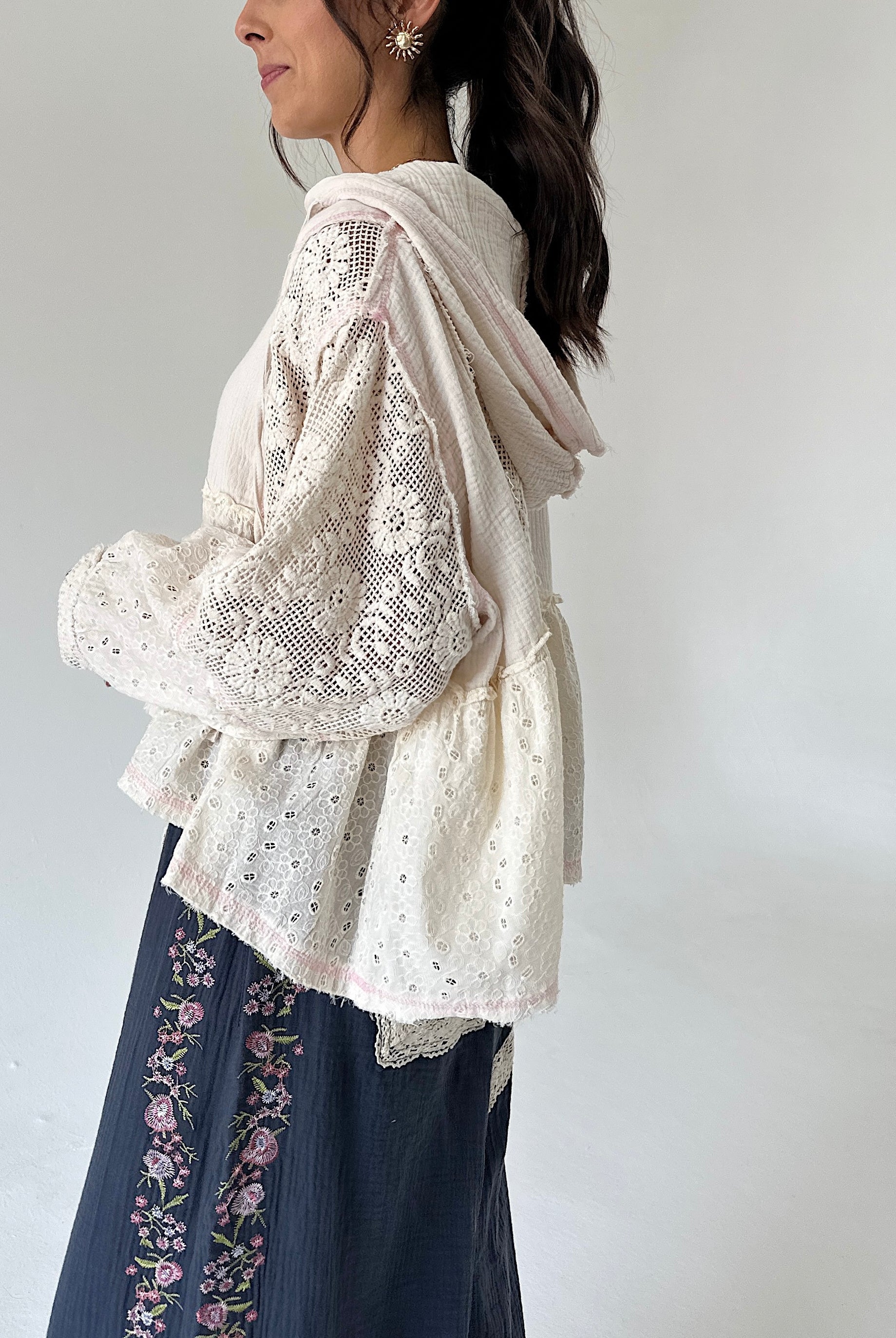 A woman shown from the side wearing a cream-colored eyelet lace long-sleeve hooded cardigan over a matching cream knit top, paired with dark blue floral embroidered pants, against a plain light gray background.