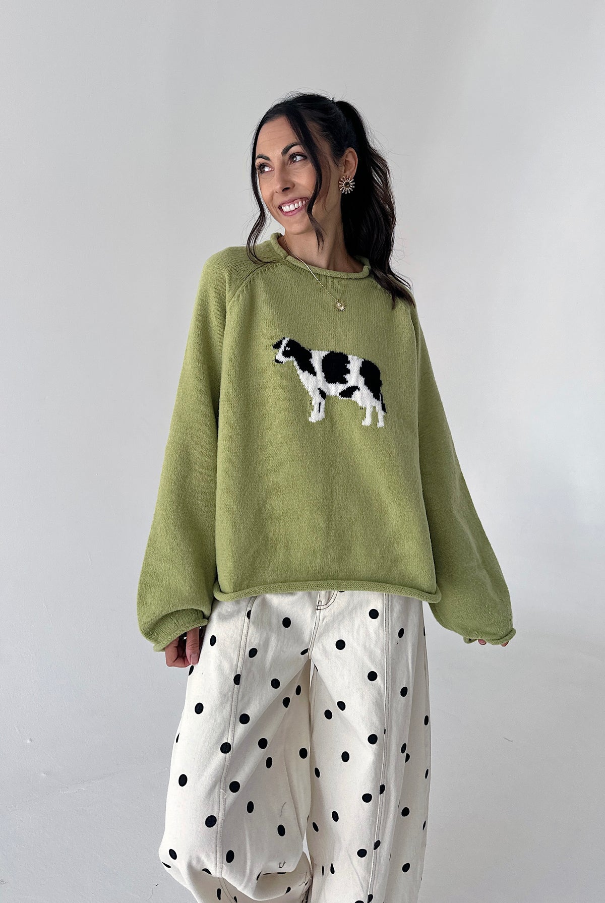 A woman wearing a sage green crewneck sweater with a black and white cow graphic on the chest, paired with white wide-leg pants featuring black polka dots, set against a plain light gray studio background.
