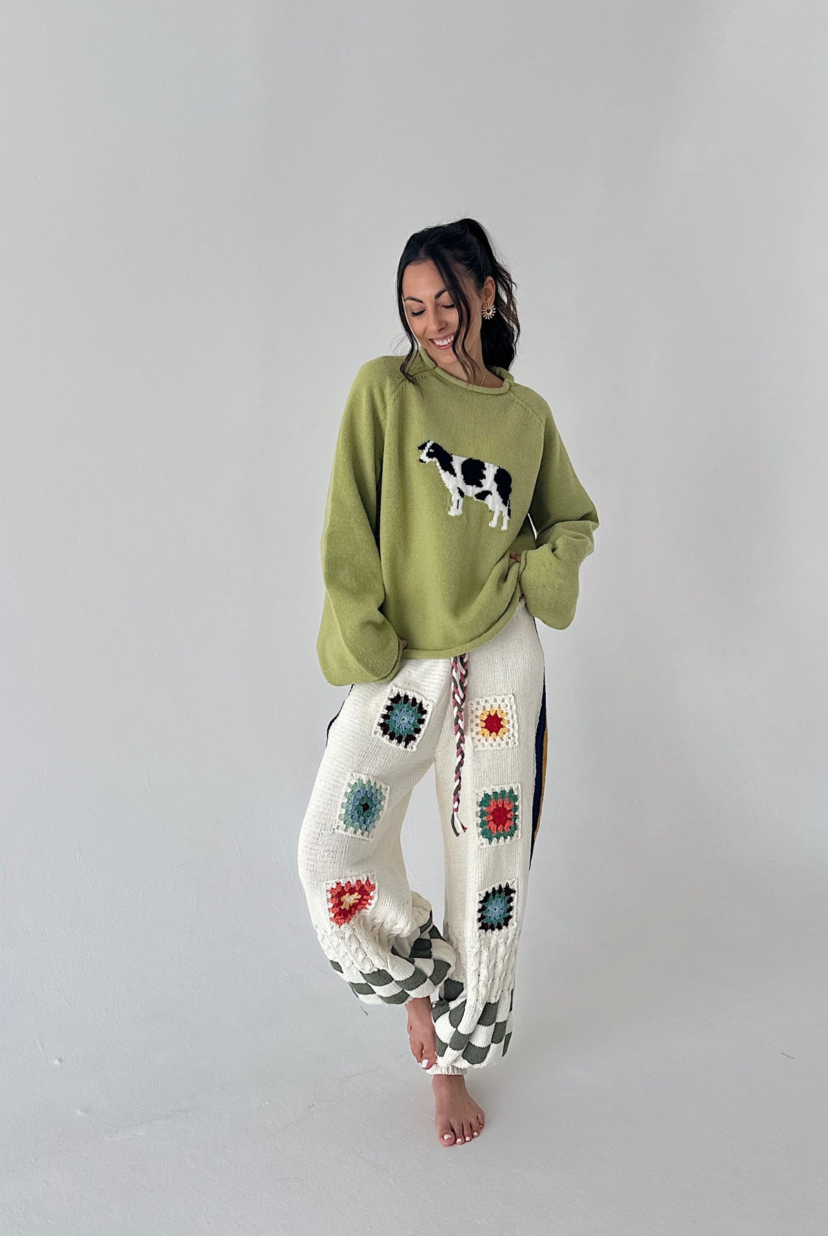 A woman stands barefoot against a plain light gray studio background, wearing a loose sage green sweater with a cow applique and white crocheted pants decorated with colorful granny-square patterns.