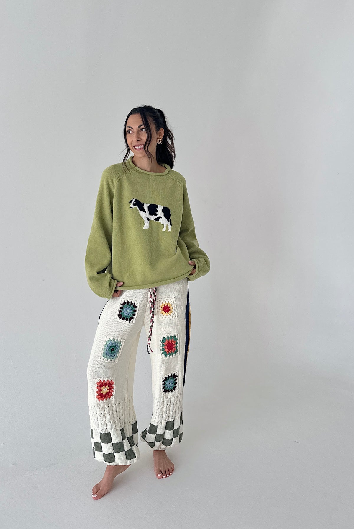 A woman stands barefoot in a studio wearing a light olive green crewneck sweater with a cow graphic and white crochet wide-leg pants with colorful floral motifs and black-and-white checkered cuffs.