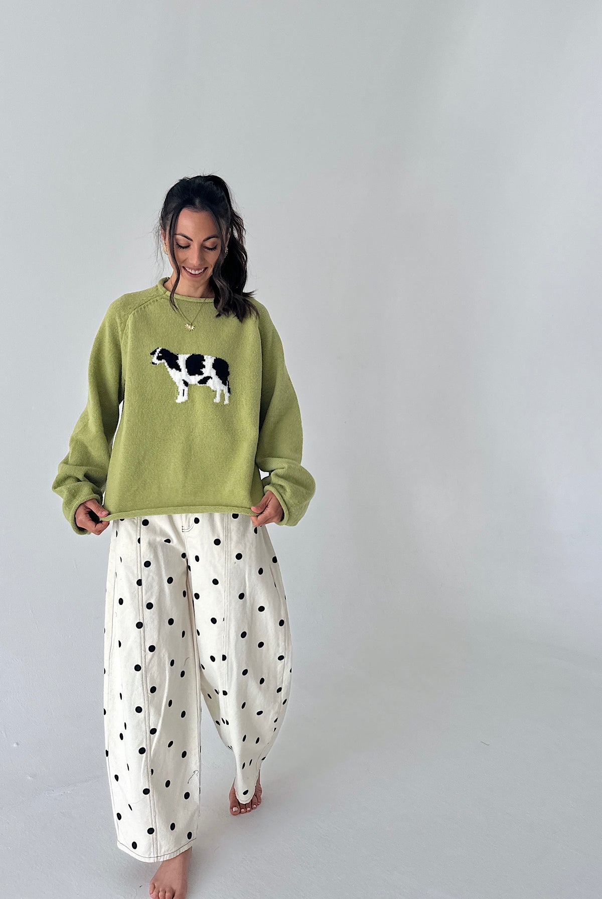 A woman modeling a light olive green crewneck sweater with a black and white cow graphic on the chest, paired with white wide-leg pants featuring black polka dots, set against a plain light gray studio background.