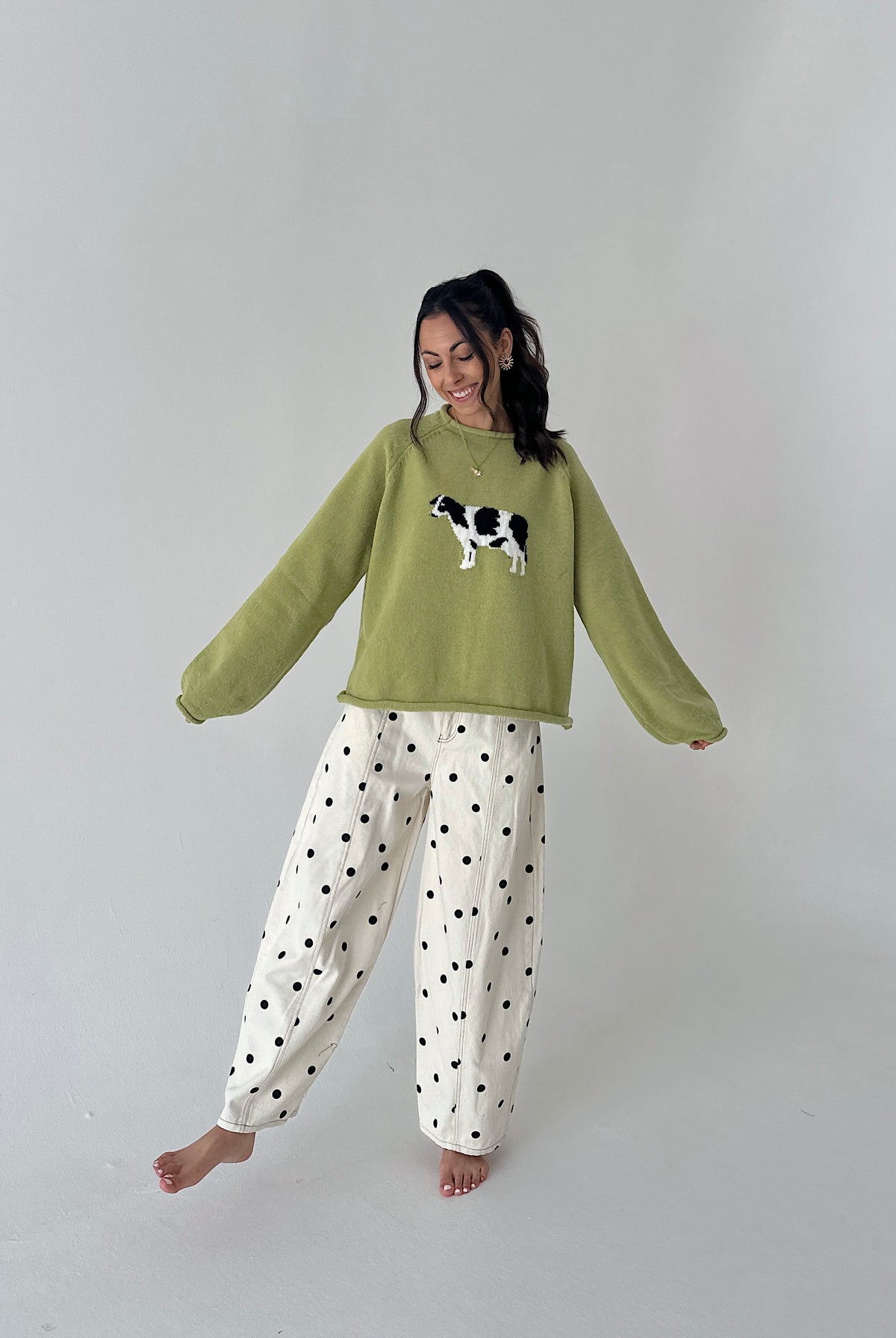 A woman modeling a cozy sage green crewneck sweater with a black and white cow applique on the chest, paired with white wide-leg pants featuring black polka dots, set against a plain light gray studio background.