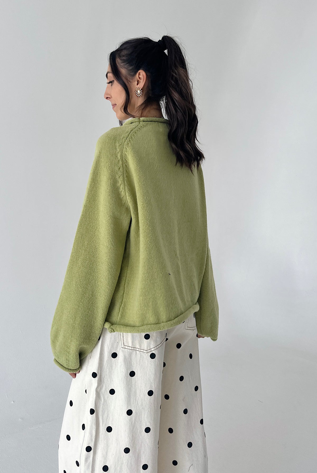 A woman shown from the back wearing a light olive green knit sweater and a white skirt with black polka dots, standing against a plain light gray wall.