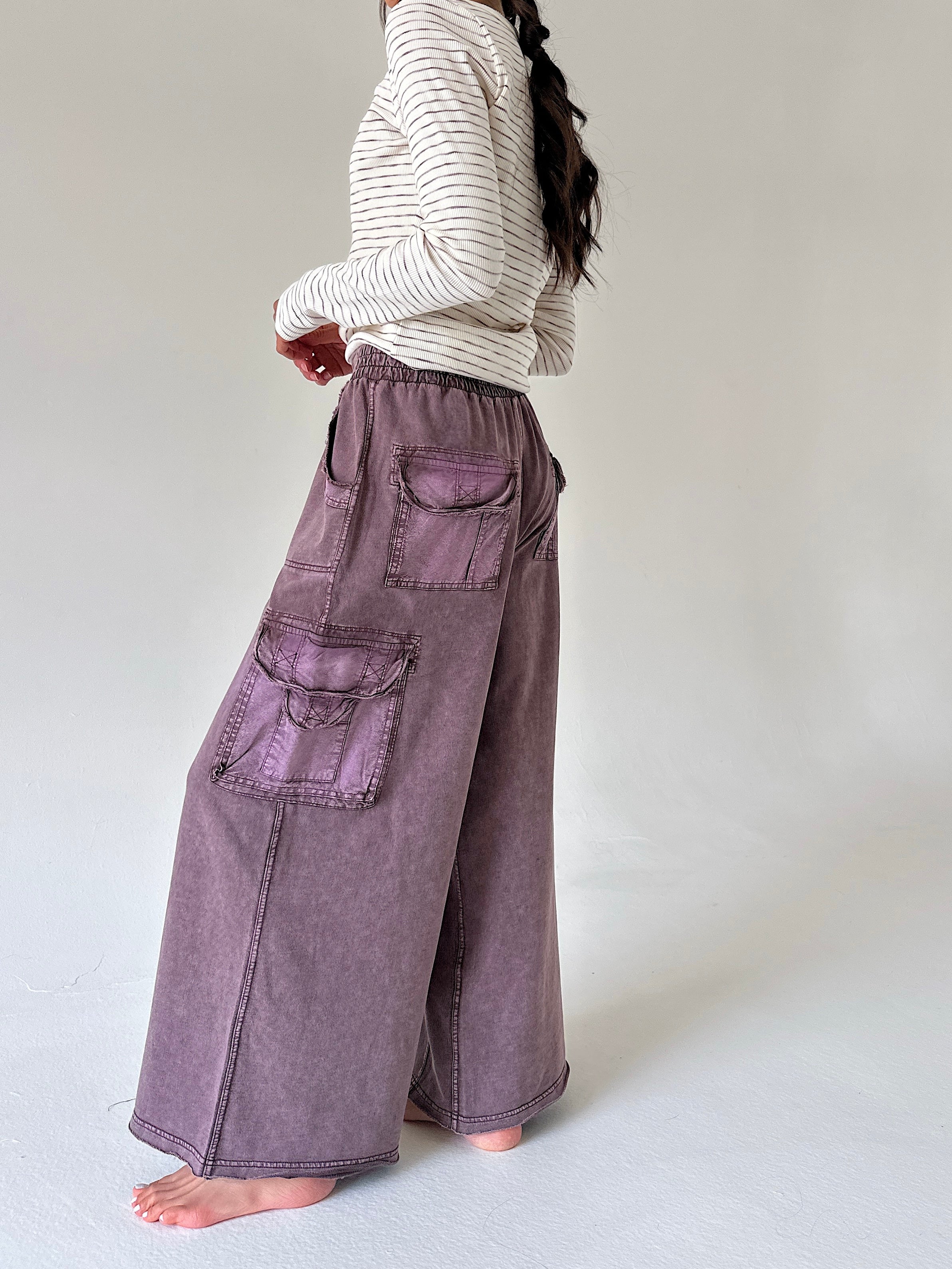 A person shown from the back wearing wide-leg mauve-purple cargo pants and a cream striped long-sleeve top, standing barefoot against a plain light gray studio background.
