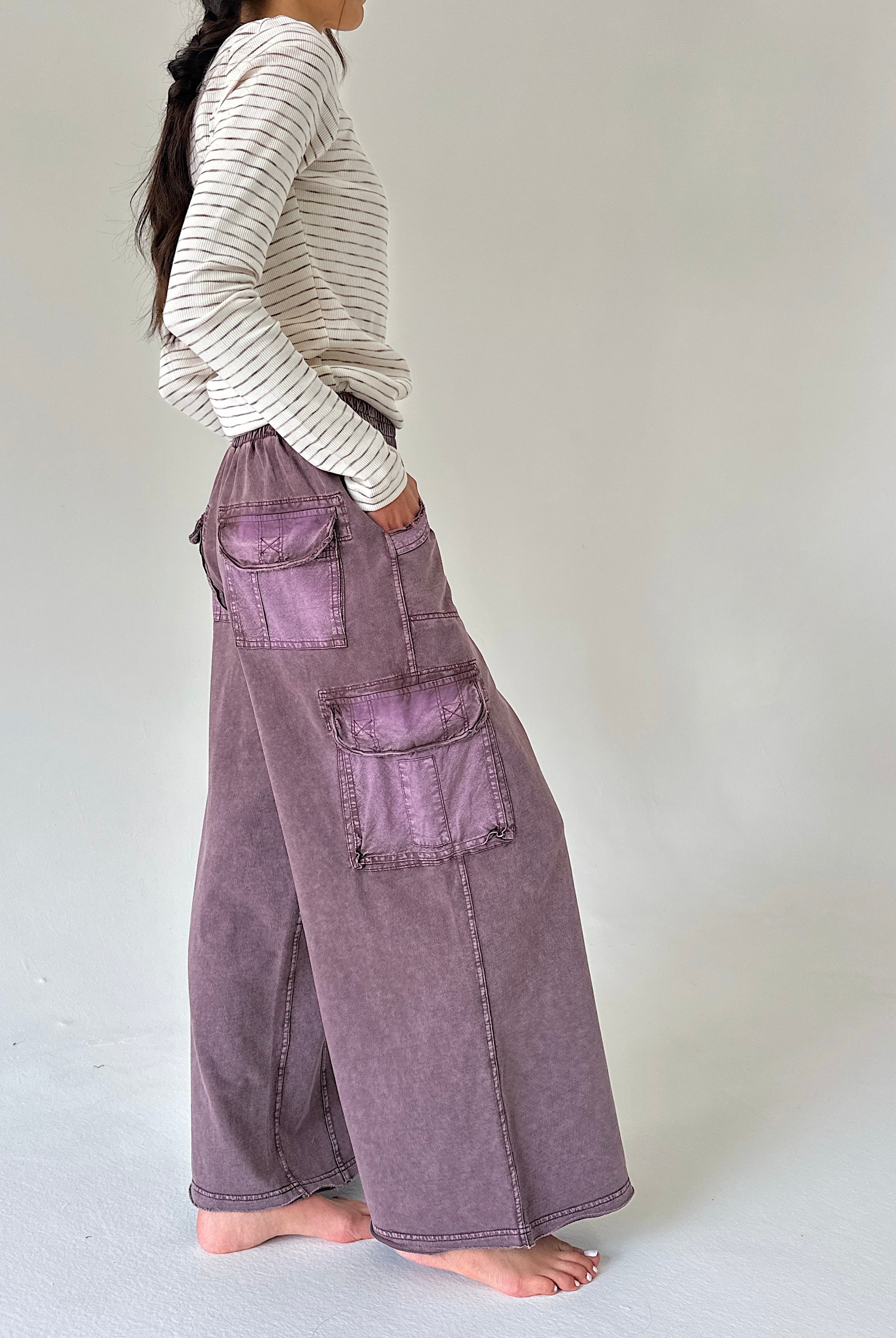 A person shown from the side wearing a cream and gray striped long-sleeve top and wide-leg mauve-purple cargo pants, standing barefoot against a plain light gray studio background.