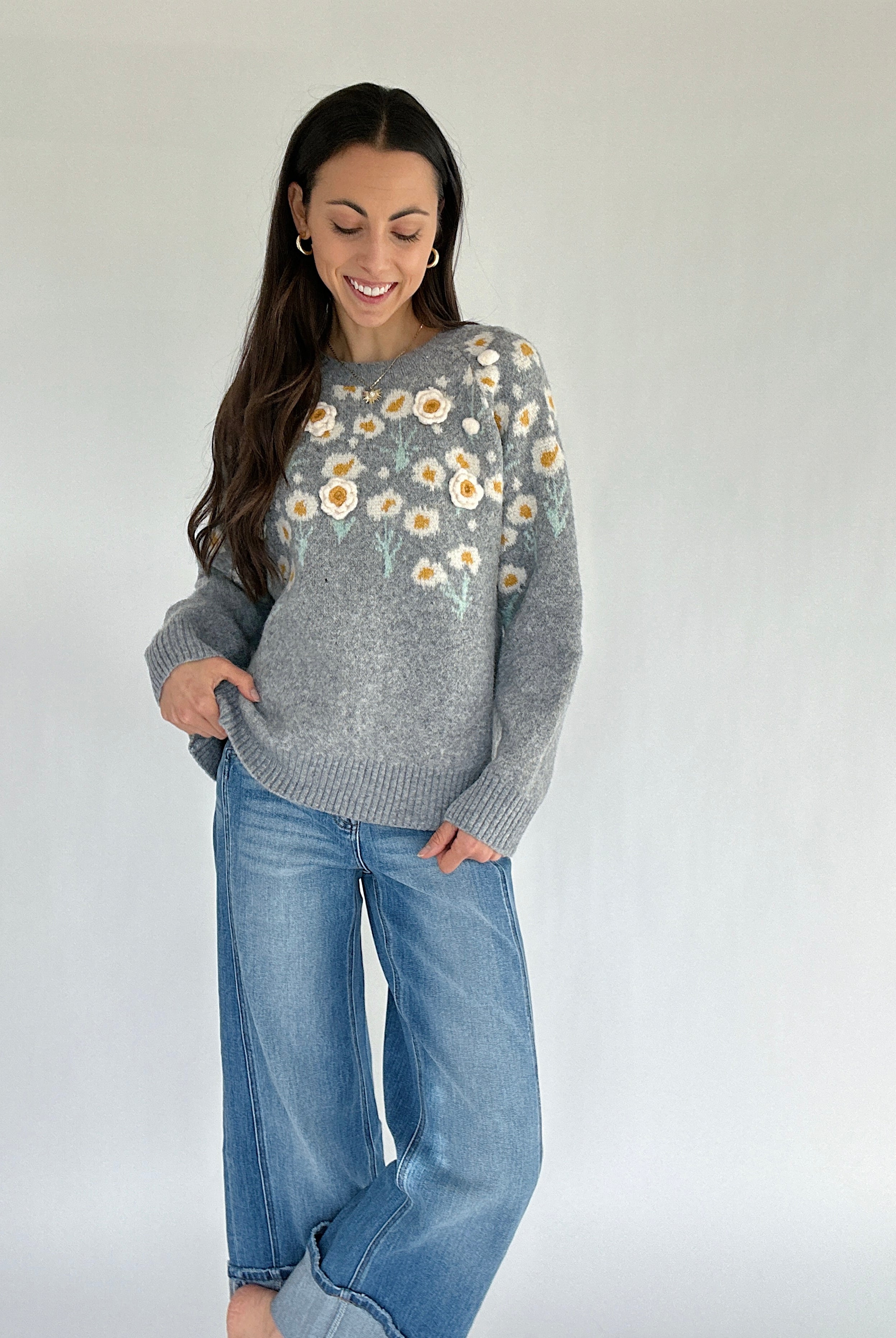 Woman wearing a gray sweater with floral patterns and blue jeans against a white background