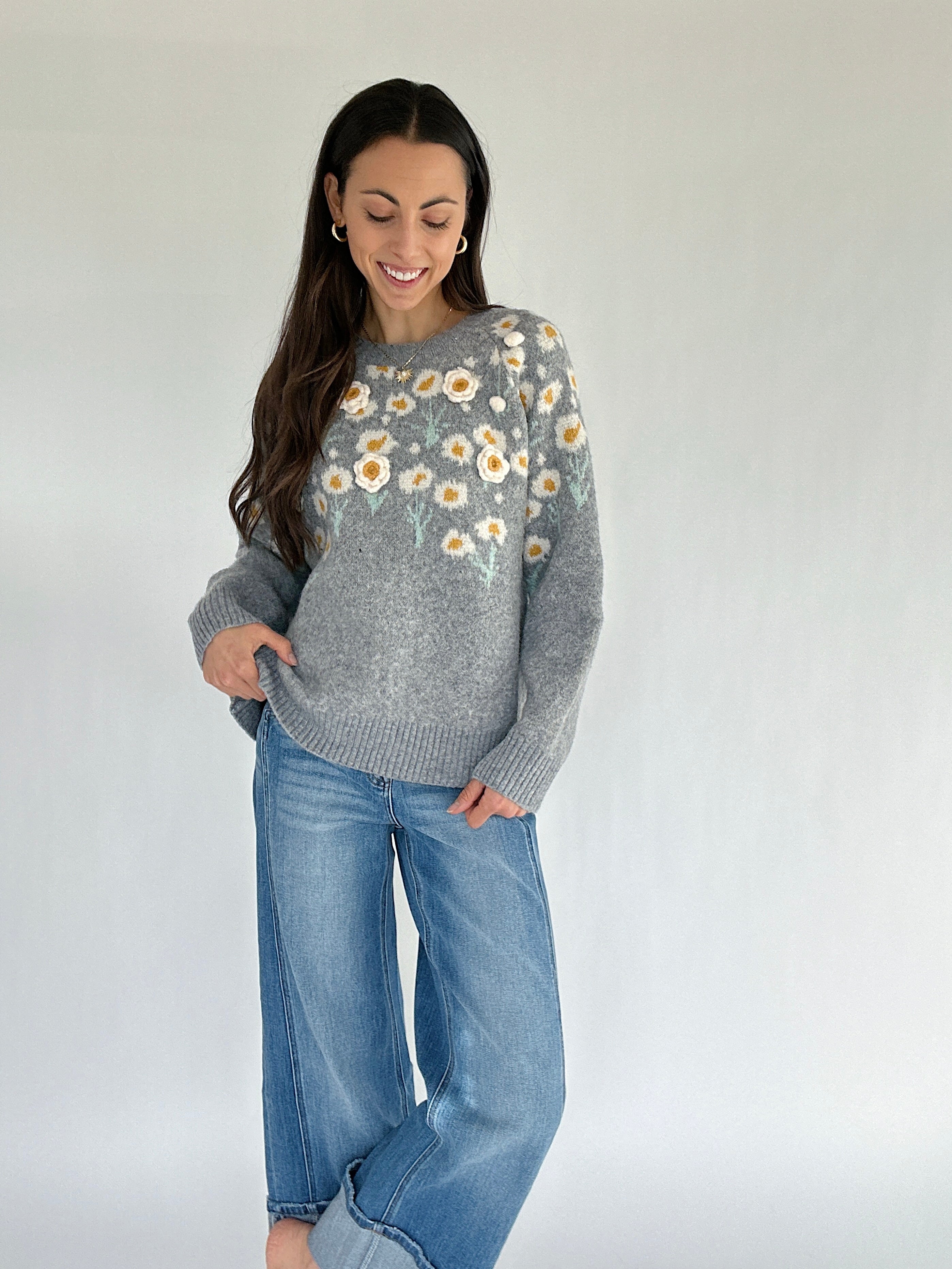 Woman wearing a gray sweater with floral patterns and blue jeans against a white background