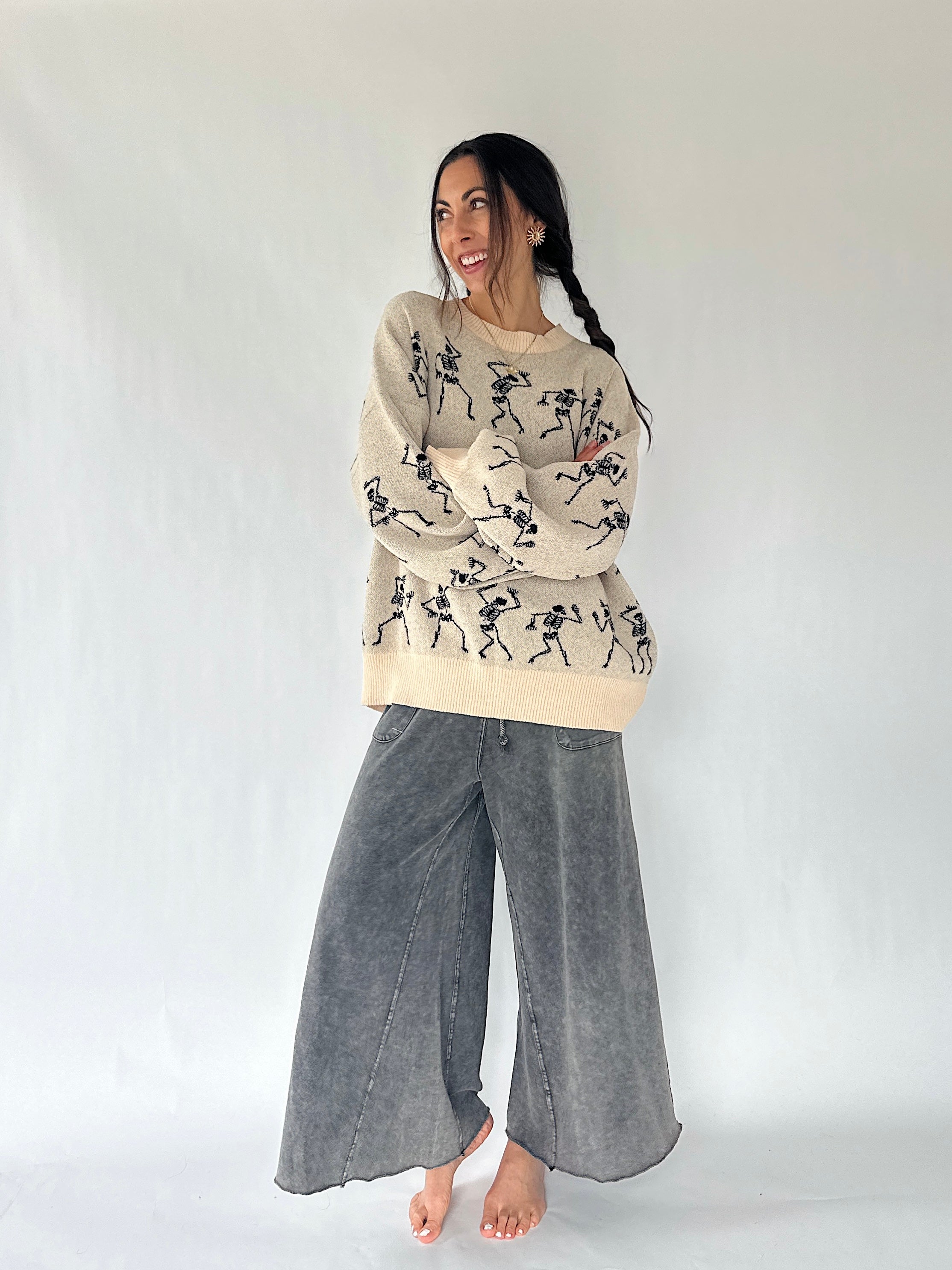 woman standing with her arms crossed wearing a cream sweater with dancing skeletons all over and a pair of wide leg sweatpants in an ash color