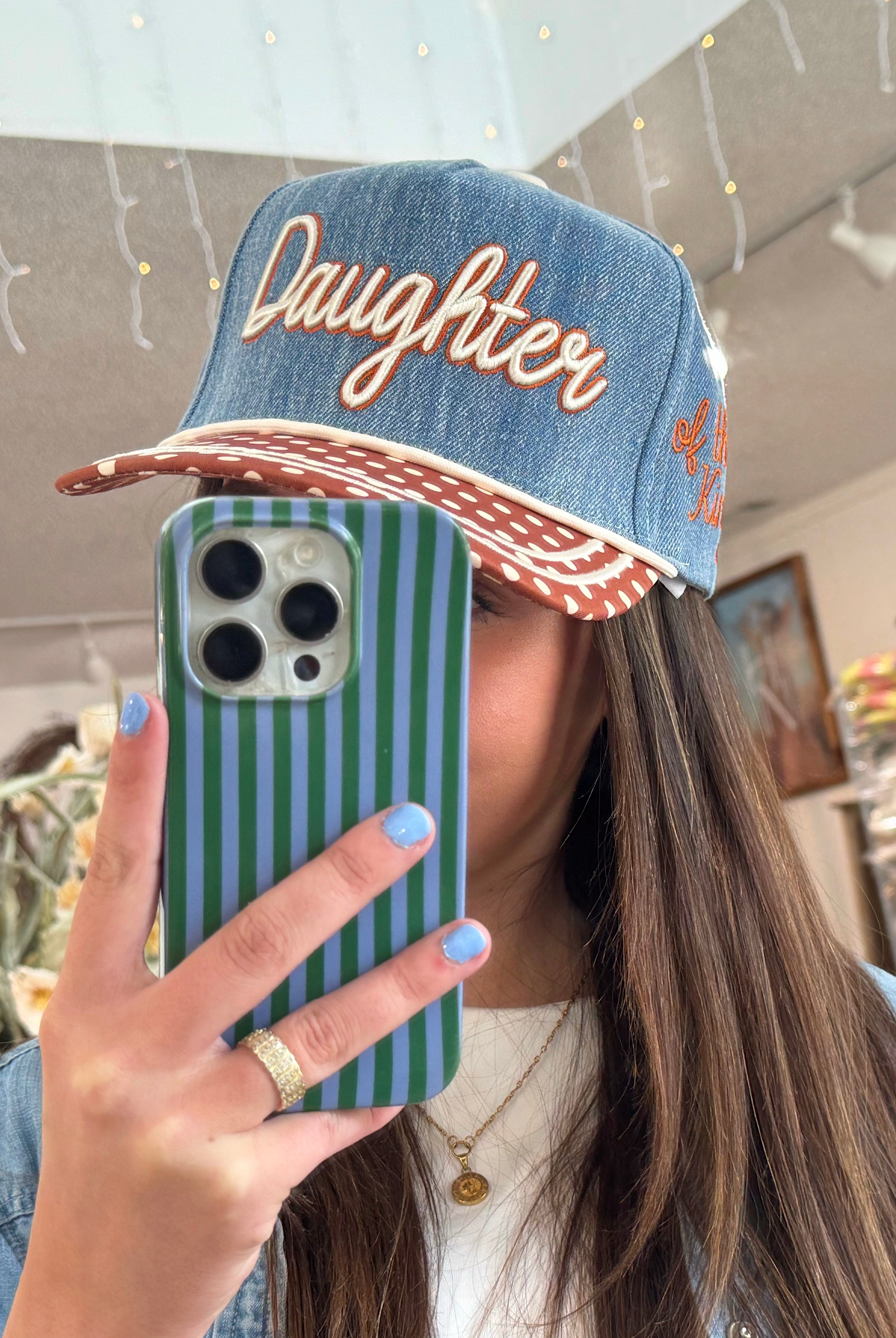Person wearing a denim cap with 'Daughter' text, holding a striped phone case.