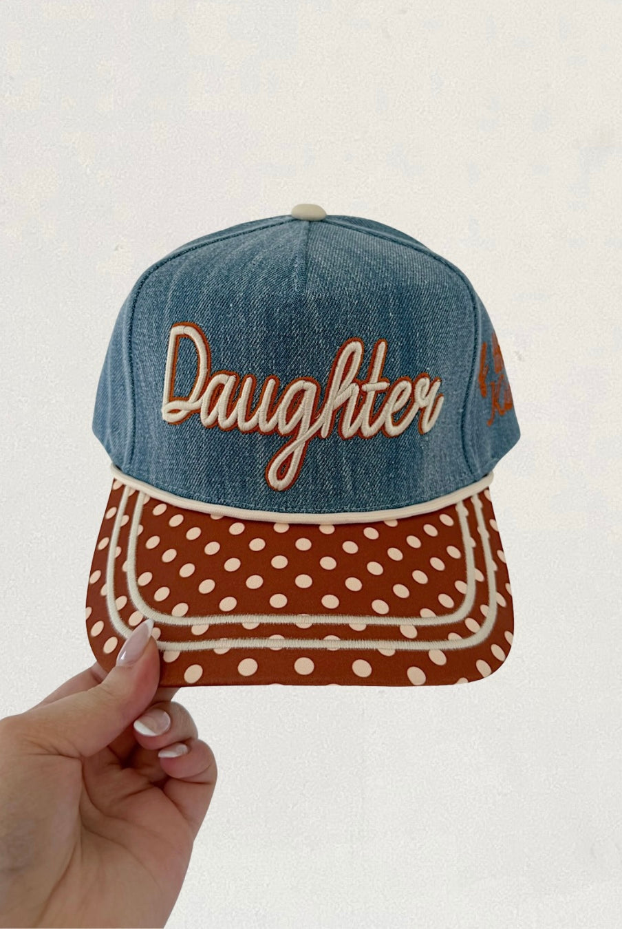 Denim cap with 'Daughter' text and polka dot pattern held by a hand against a light background