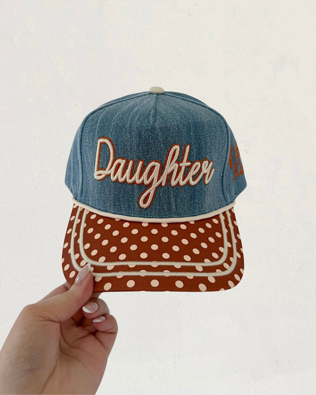 Denim cap with 'Daughter' text and polka dot pattern held by a hand against a light background