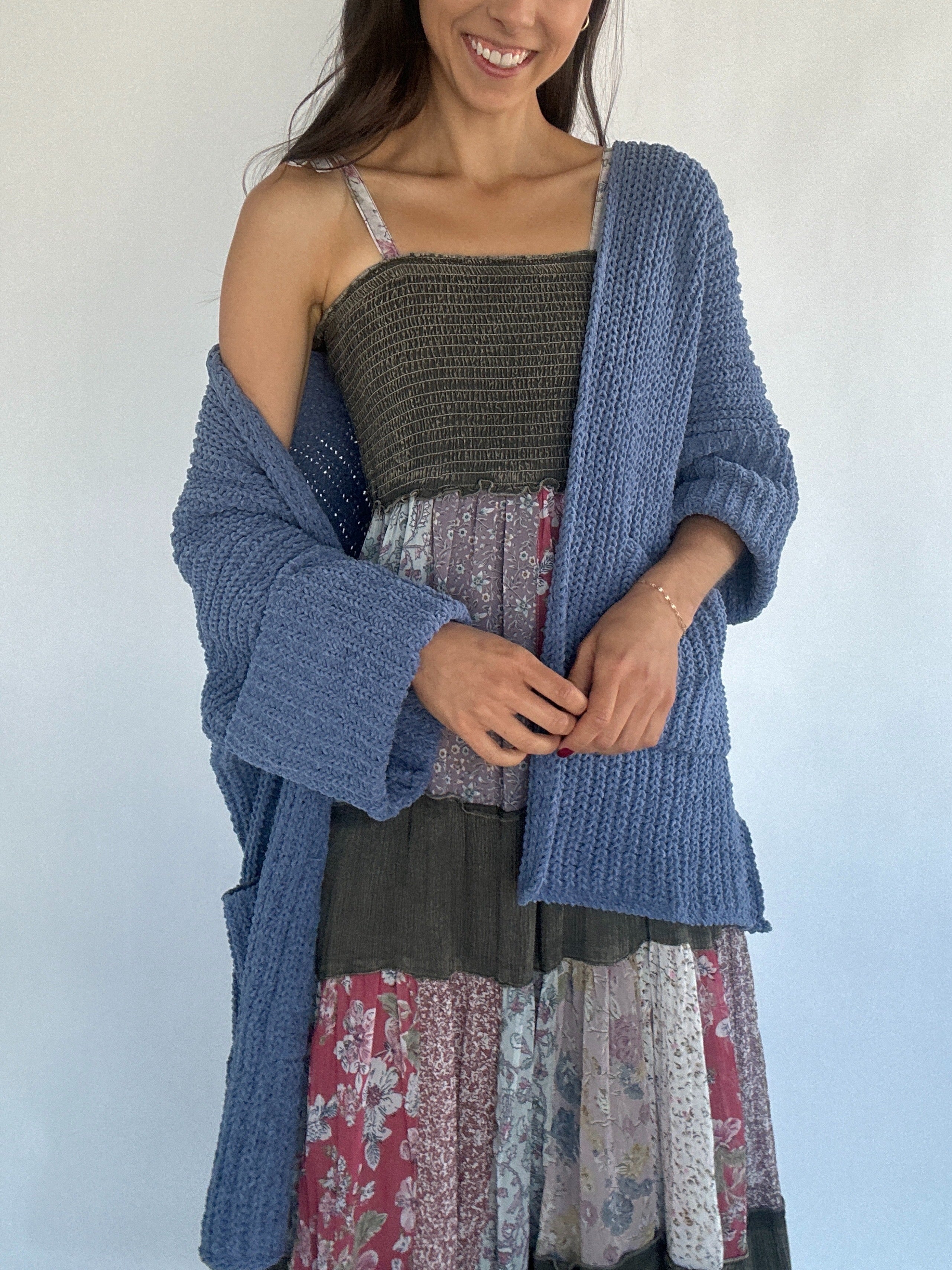 Woman wearing a blue chenille knitted cardigan off one shoulder over a patterned dress against a white background