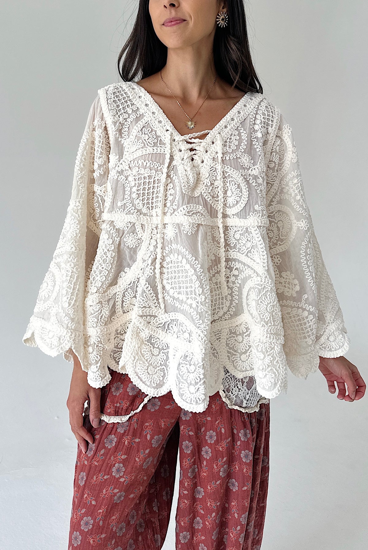A woman wearing a cream-colored, long-sleeved lace top with a lace-up neckline and scalloped edges, paired with rust-colored floral pants, against a plain light gray background.