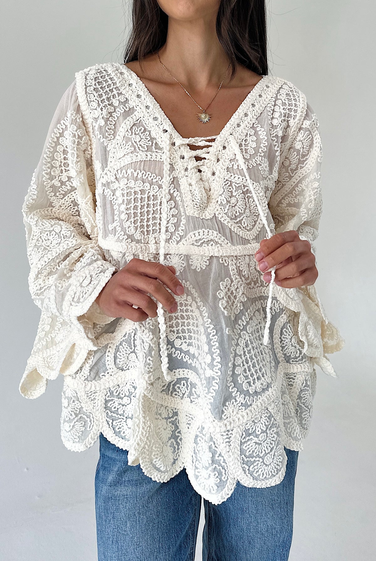 A woman wearing a cream-colored lace blouse with long bell sleeves, a V-neckline with tie-front details, and a scalloped hem, paired with blue jeans against a plain light gray background.