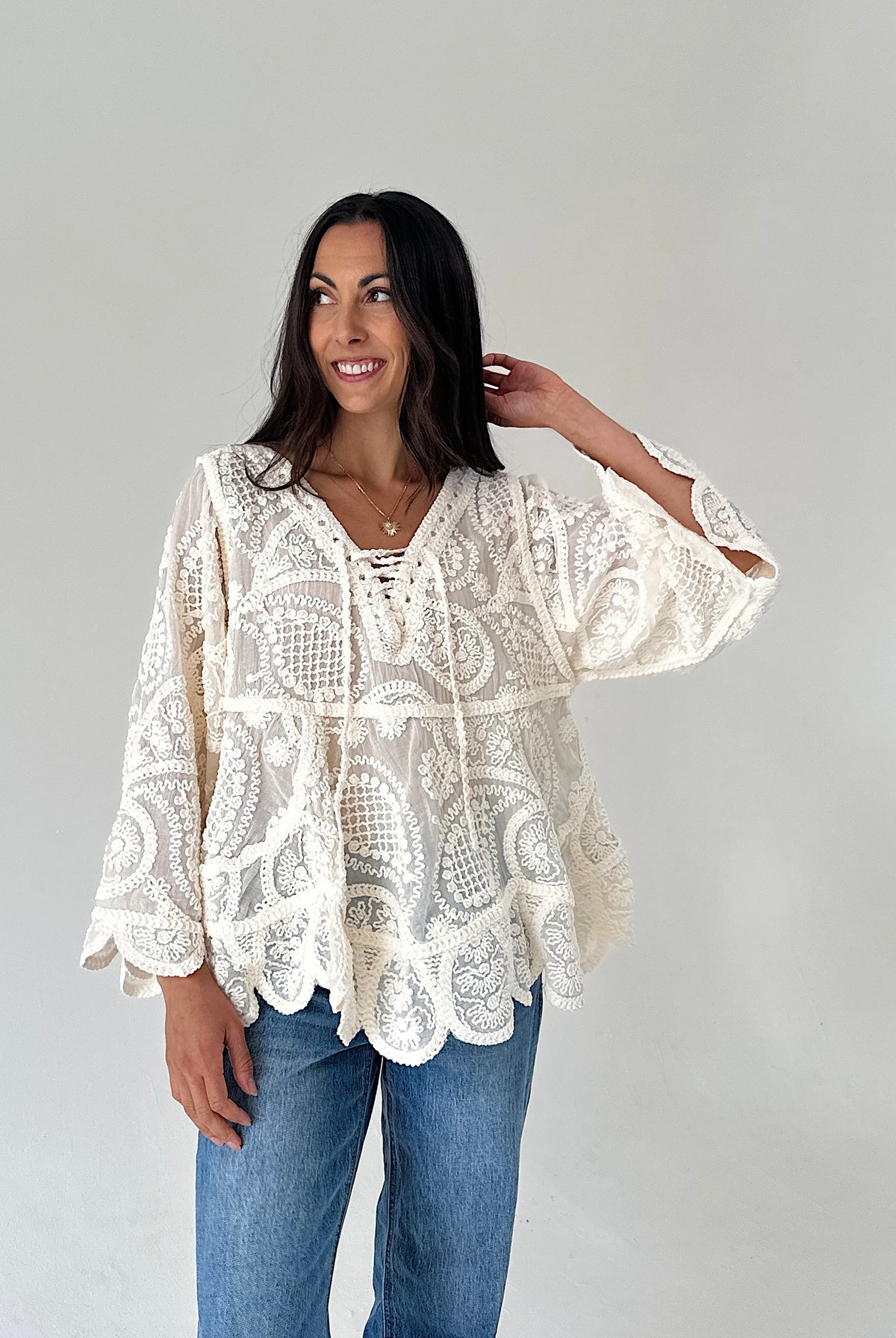 A woman modeling a cream lace bohemian-style top with blue jeans, standing against a plain light gray studio background.