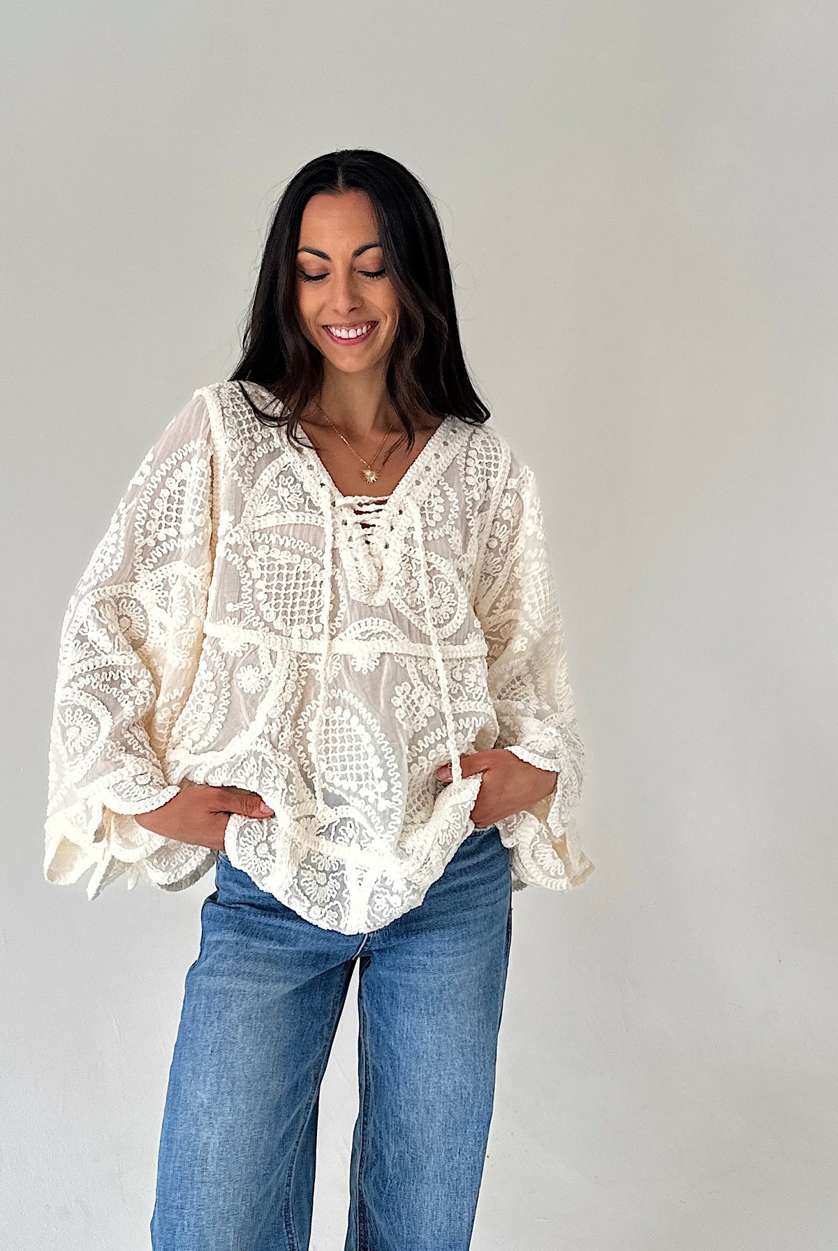 A woman wearing a cream-colored crochet lace long-sleeve blouse with blue jeans, standing against a plain light gray background.