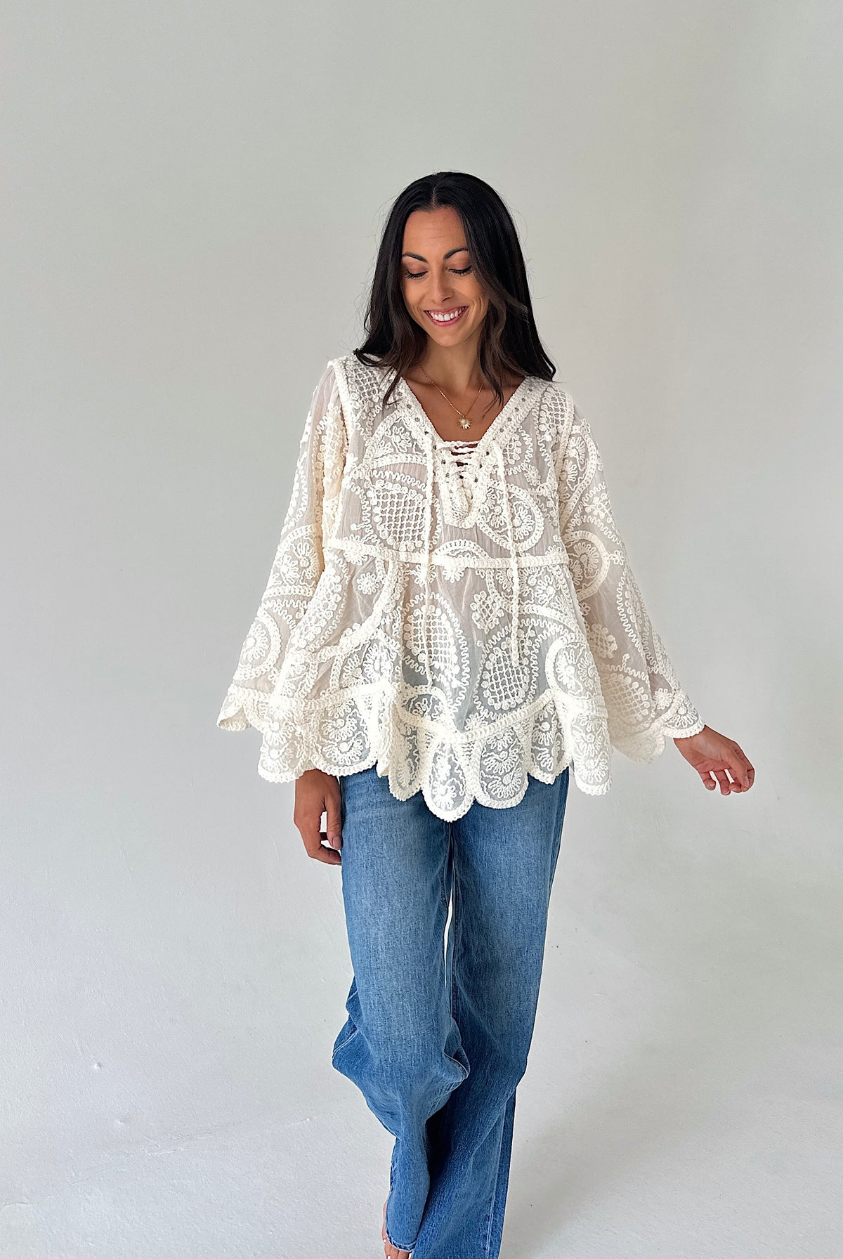 A woman wearing a cream lace bohemian-style long-sleeve top and blue jeans, standing against a plain light gray studio background.