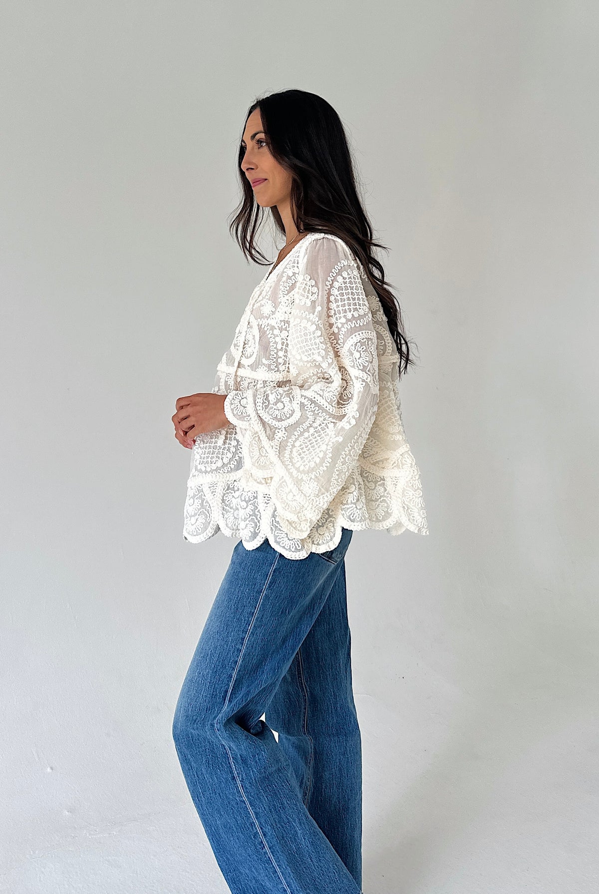 A woman wearing a cream-colored, long-sleeved lace blouse and wide-leg blue jeans, standing in profile against a plain light gray studio background.