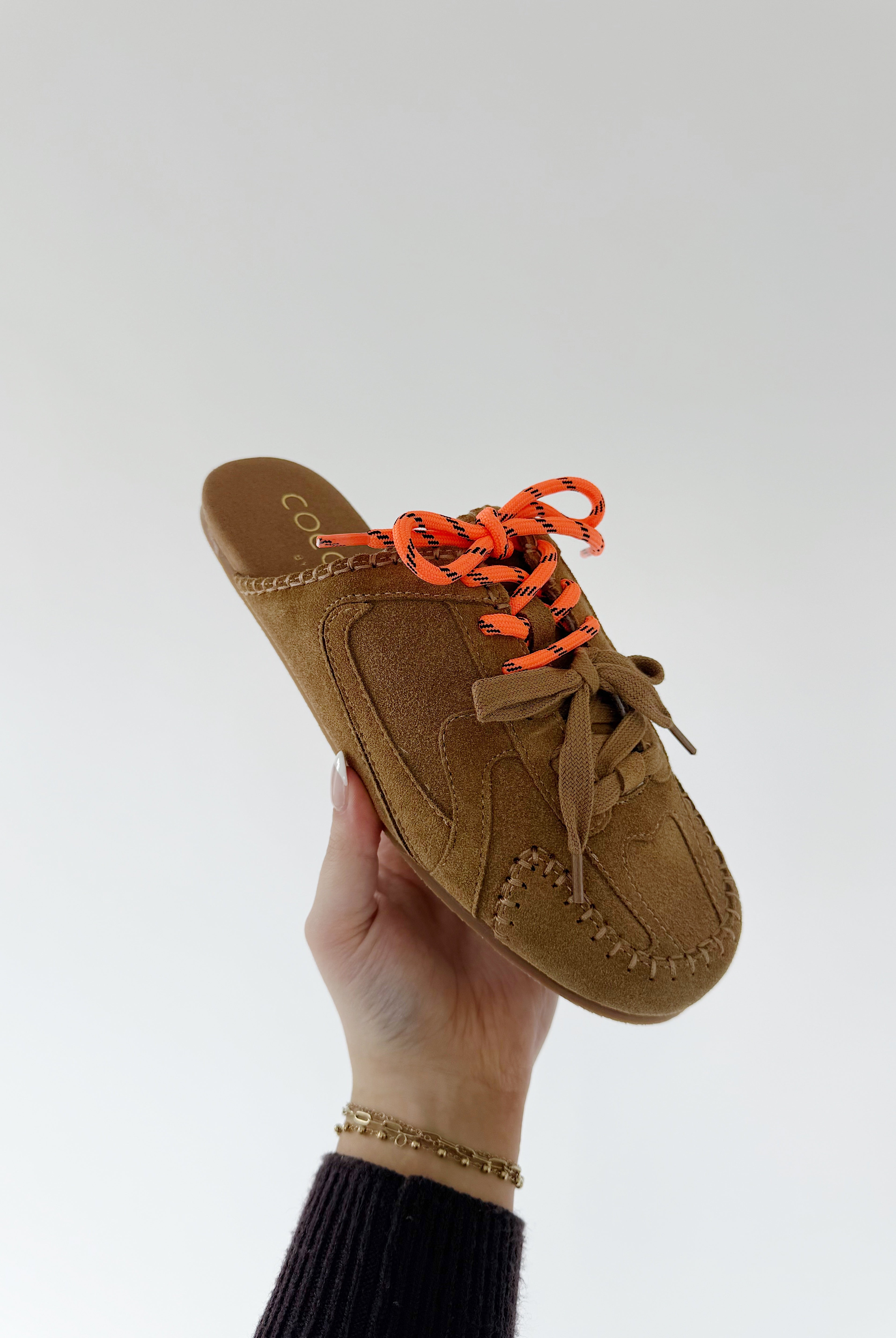 Brown slip-on shoe with orange laces held by a hand against a white background