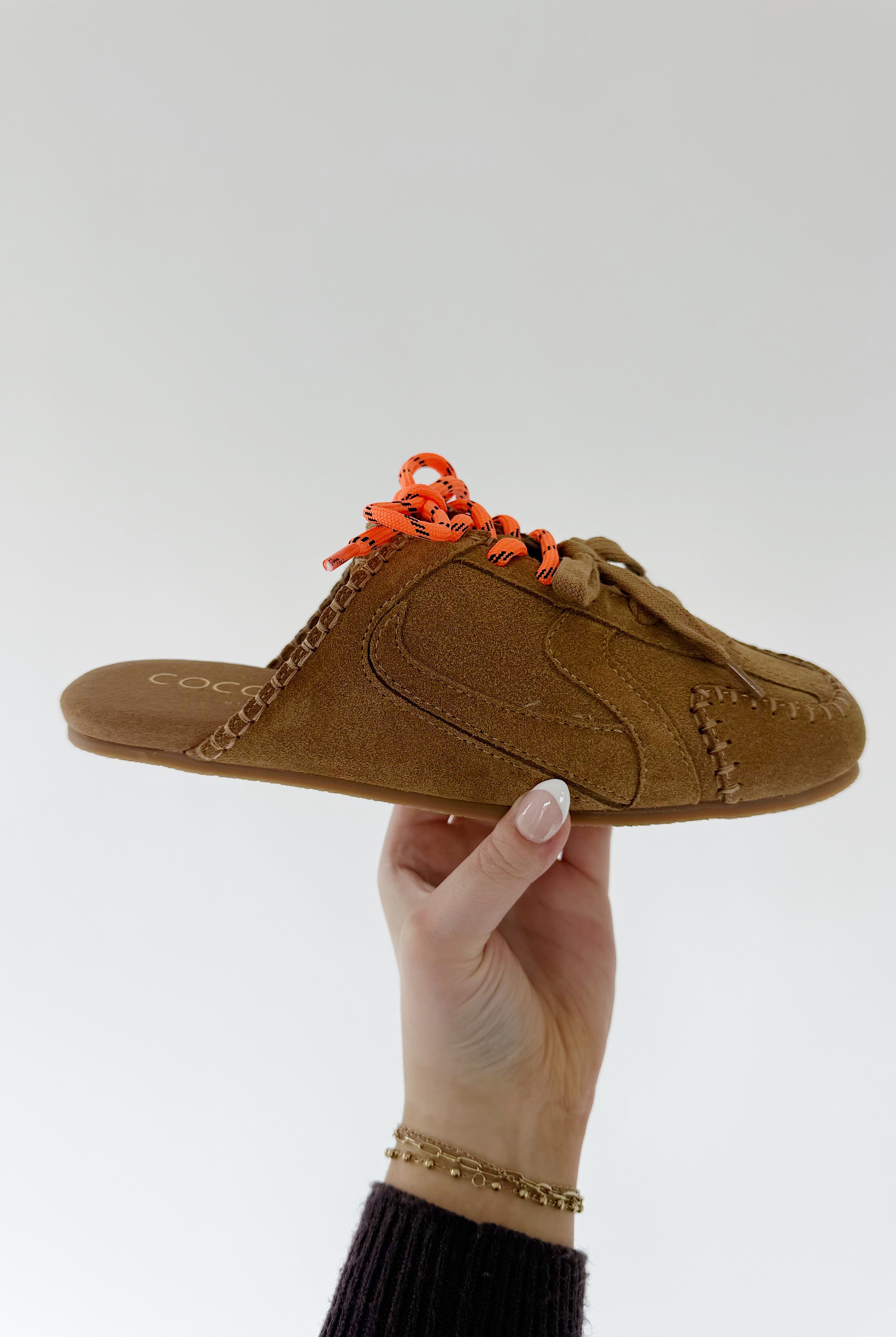 Brown sneaker mule with orange laces held by a hand against a white background