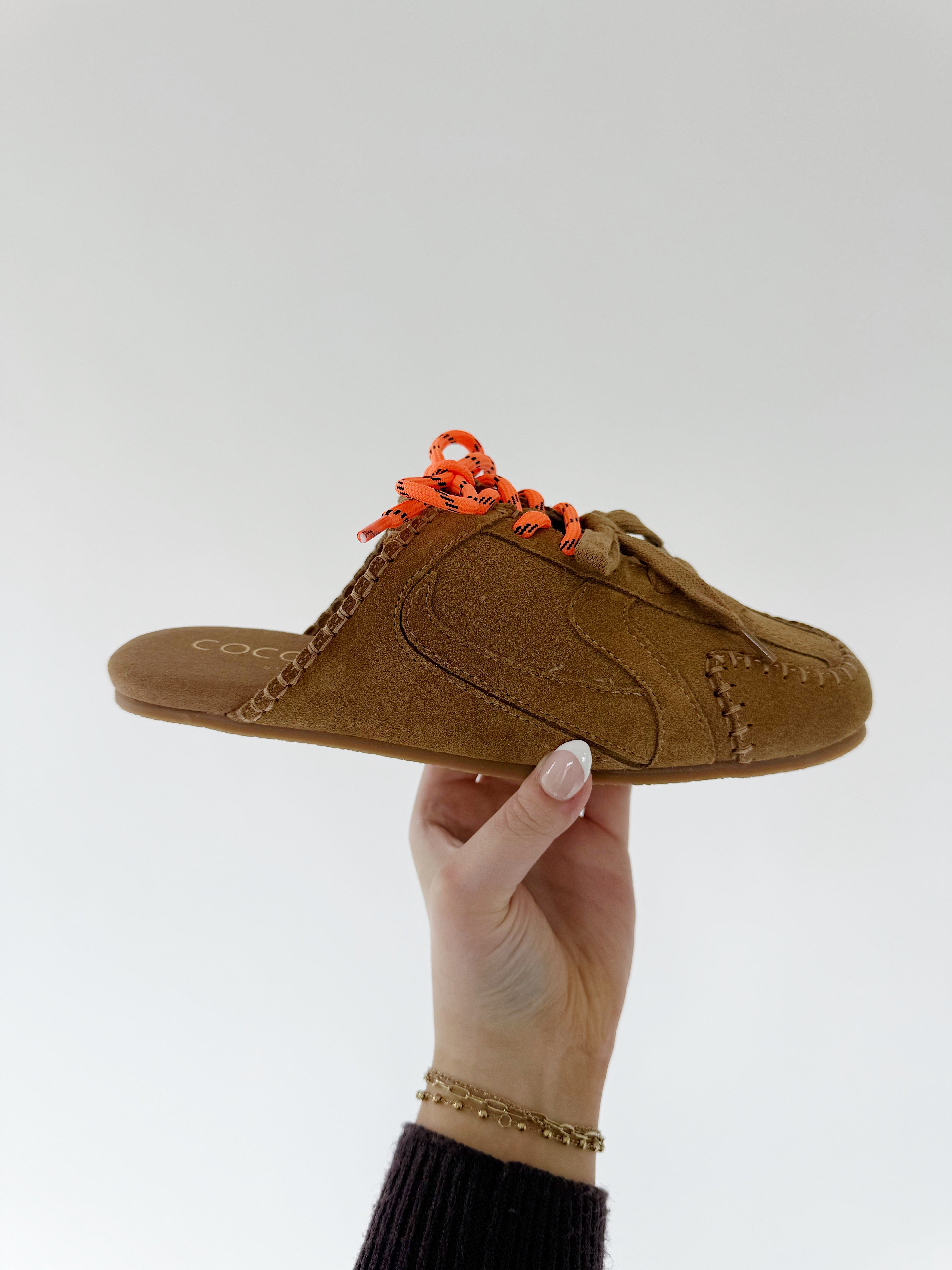 Brown sneaker mule with orange laces held by a hand against a white background