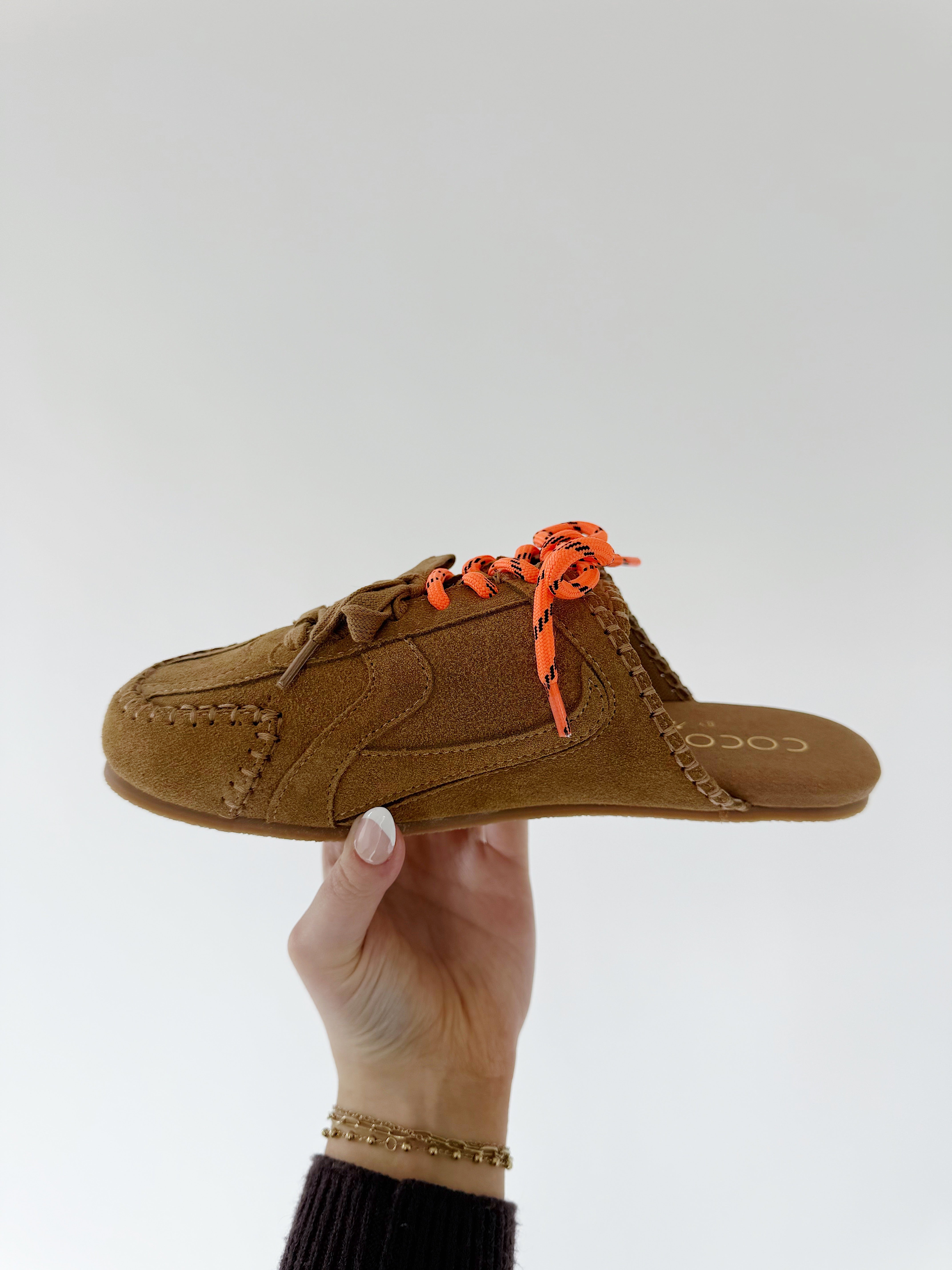 Brown slip-on shoe with orange laces held by a hand against a white background from side