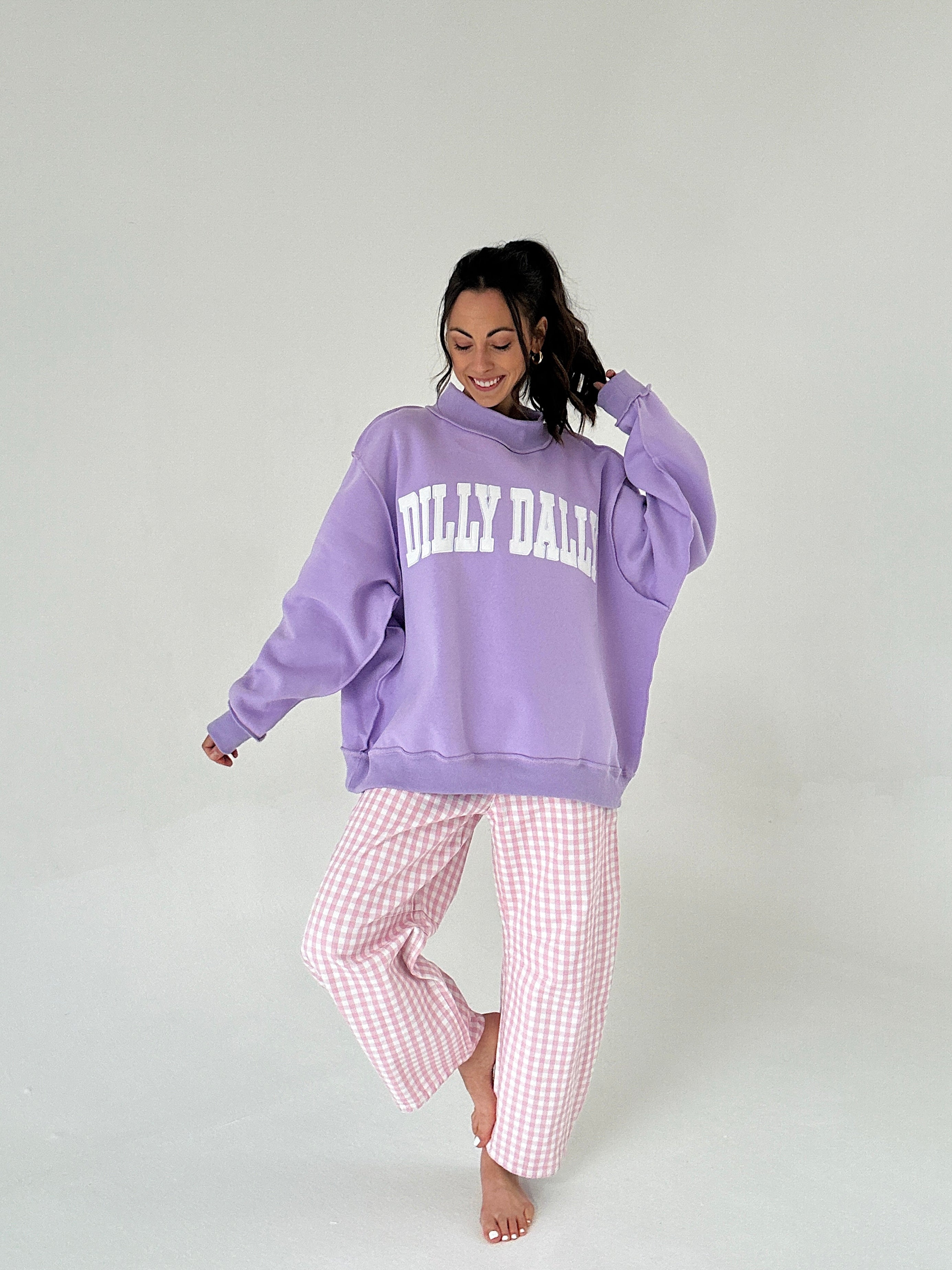 Woman wearing a purple 'Dilly Dally' hoodie and pink checkered pants on a white background