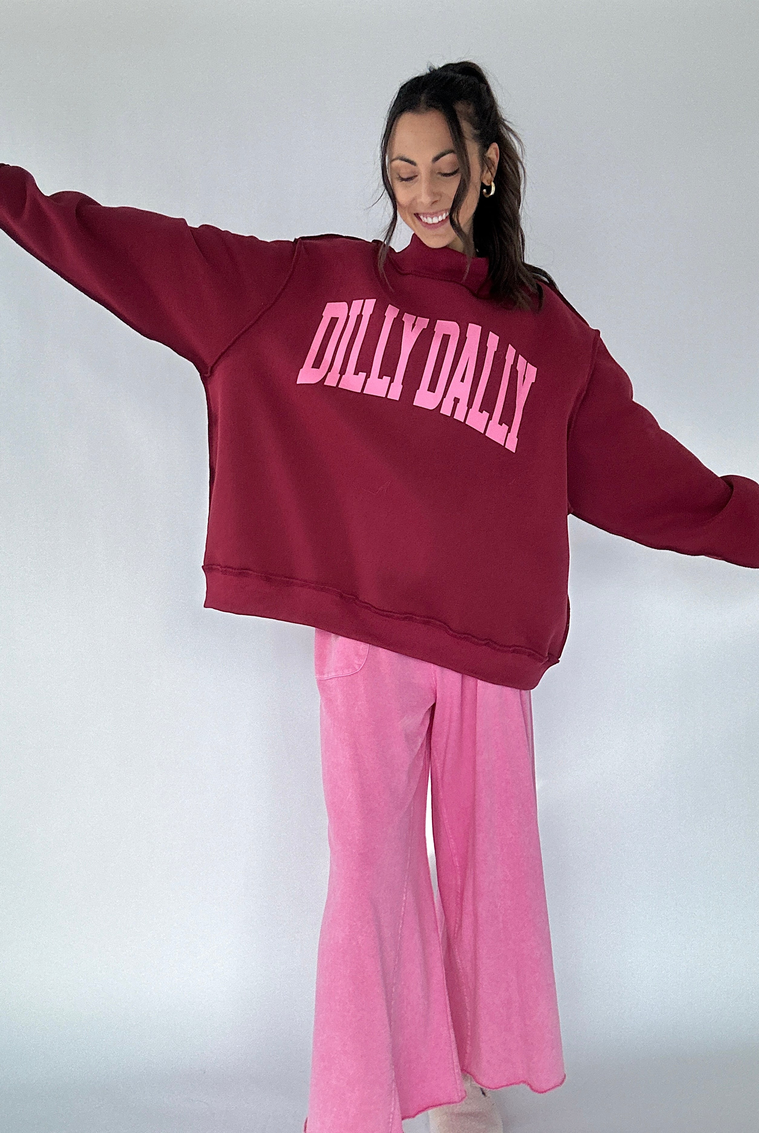 Person wearing a maroon mockneck pullover with pink 'DILLY DALLY' text and pink pants on a white background