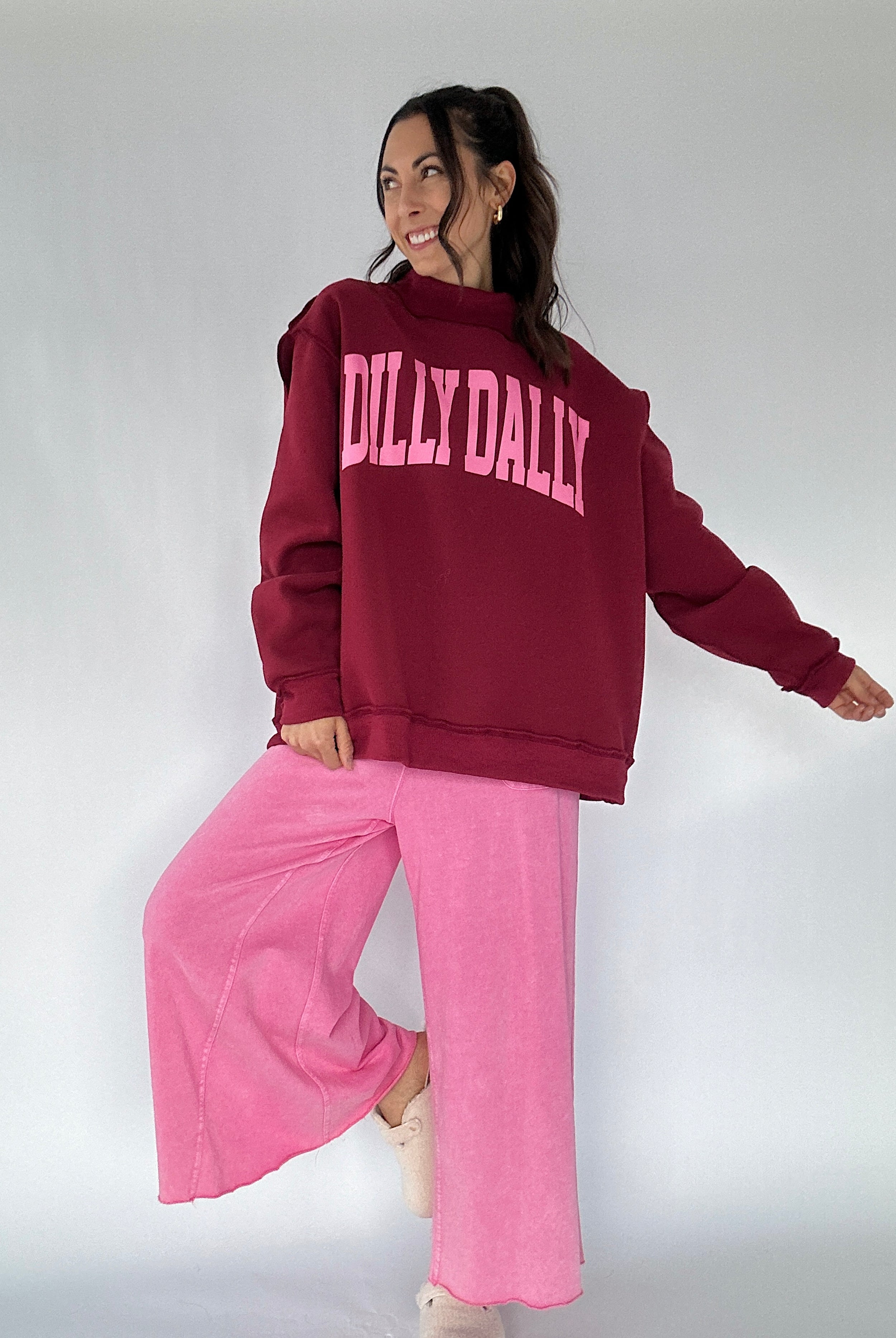 Person wearing a maroon 'DILLY DALLY' sweatshirt and pink pants on a white background