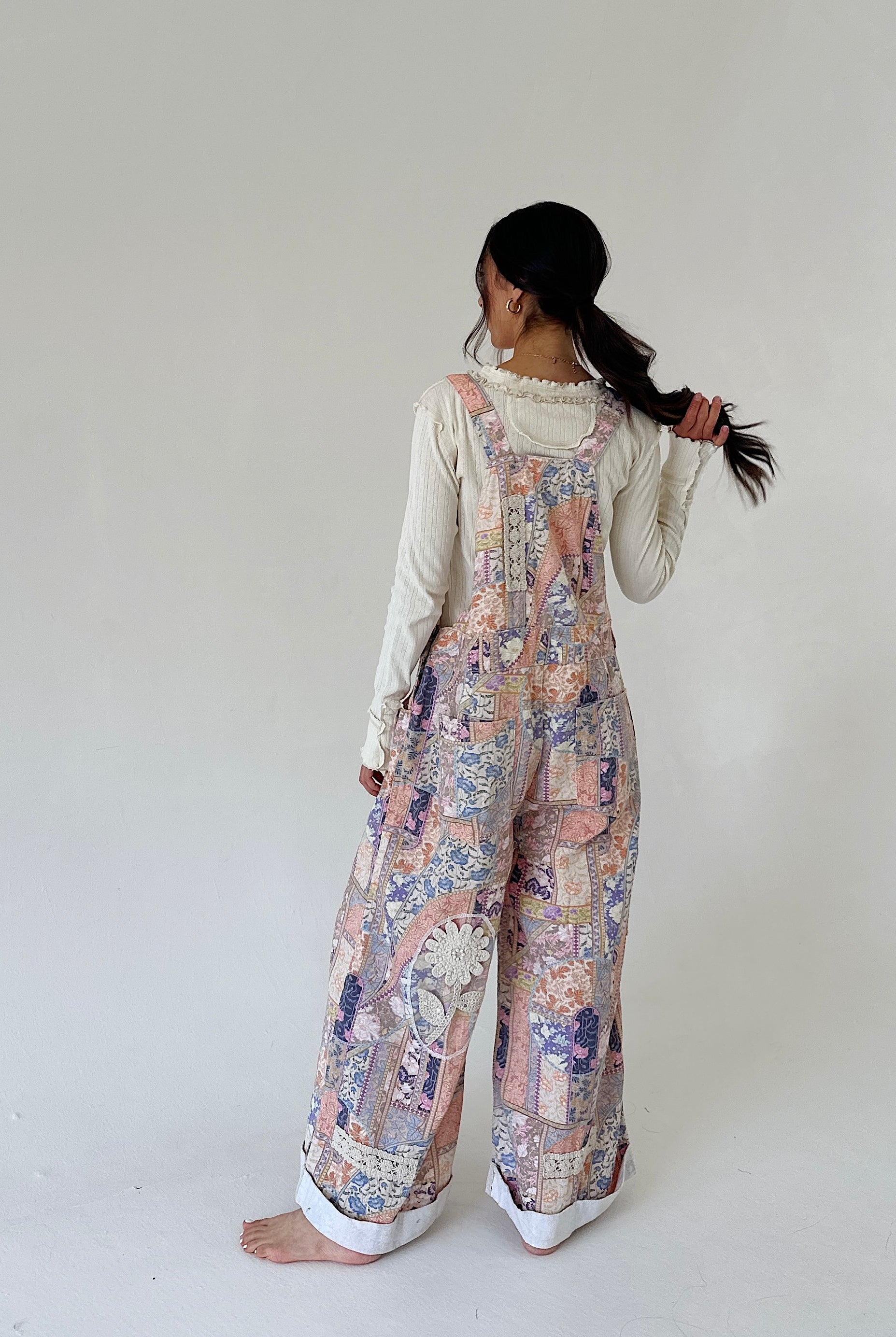 A woman stands barefoot against a plain off-white studio background, photographed from the back. She is wearing a long-sleeve cream top under loose, wide-leg overalls with a pastel multicolor patchwork print in pink, blue, beige, and purple tones.