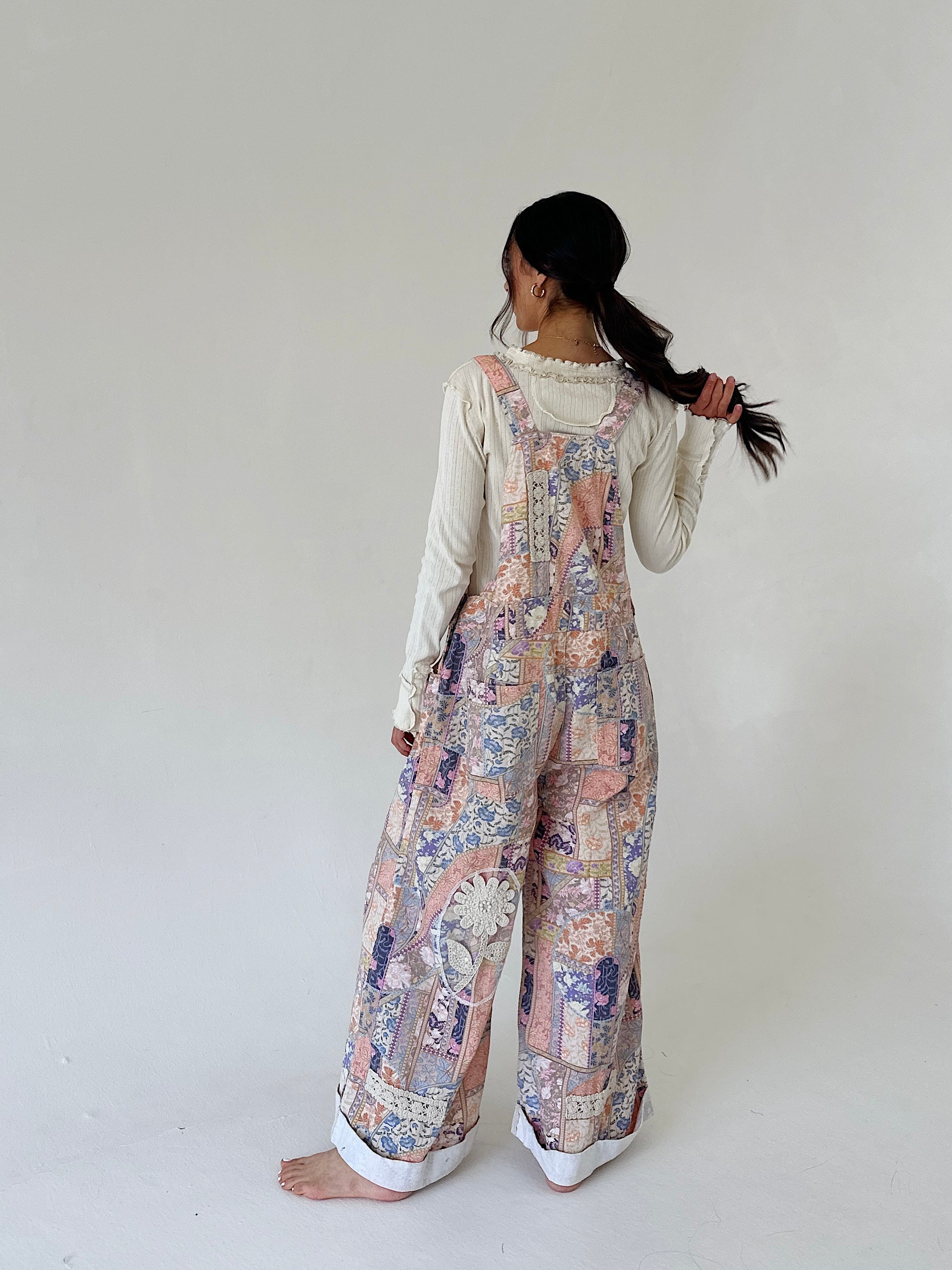 A woman stands barefoot against a plain off-white studio background, photographed from the back. She is wearing a long-sleeve cream top under loose, wide-leg overalls with a pastel multicolor patchwork print in pink, blue, beige, and purple tones.