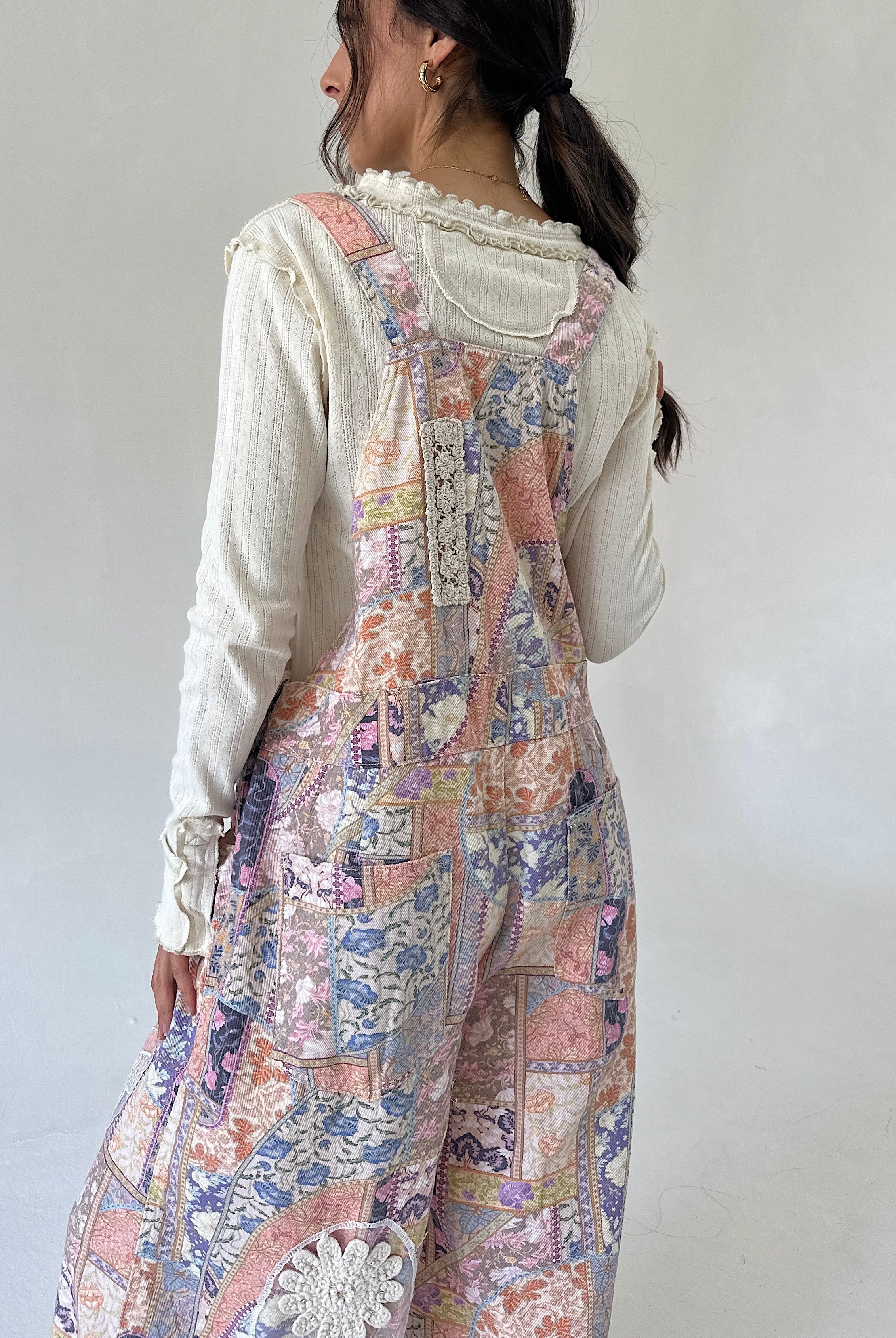 A woman shown from the back wearing a cream ribbed long-sleeve top layered under pastel patchwork overalls with floral and paisley prints in pink, blue, and beige.