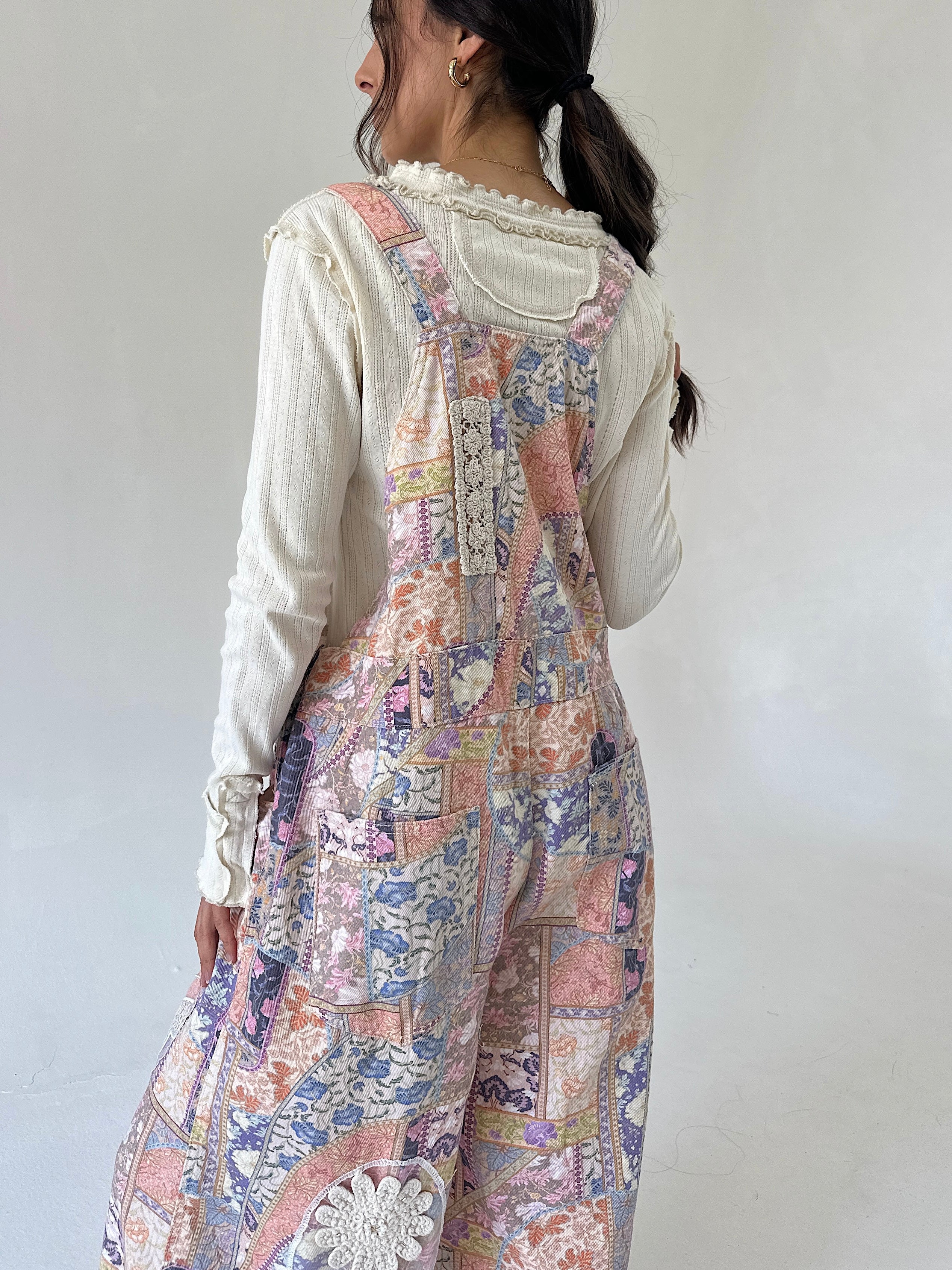 A woman shown from the back wearing a cream ribbed long-sleeve top layered under pastel patchwork overalls with floral and paisley prints in pink, blue, and beige.