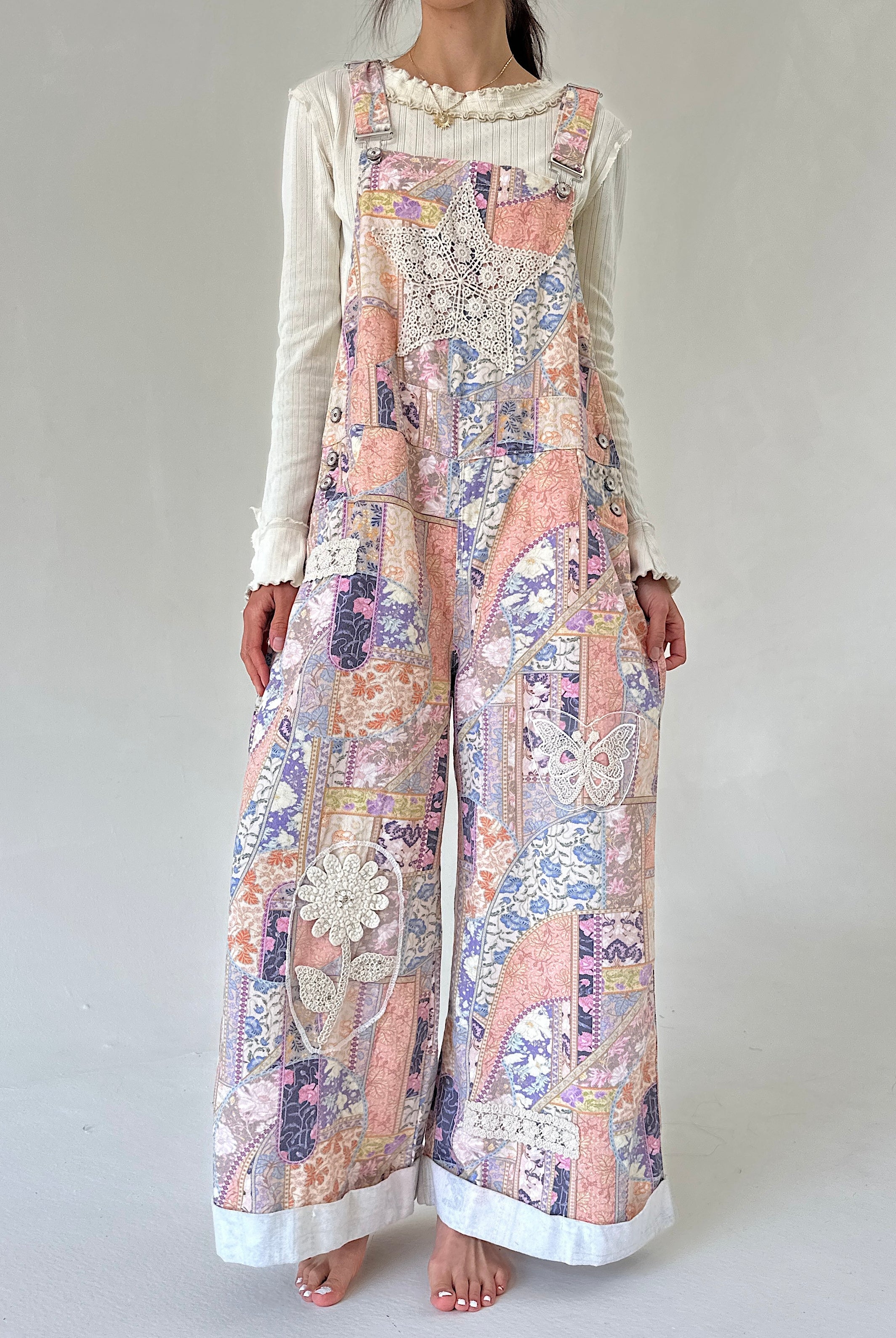 A woman modeling a lwide-leg overall jumpsuit with a multicolor patchwork print in pastel pink, purple, blue, and beige, layered over a white long-sleeve ruffled shirt, against a plain light gray background.