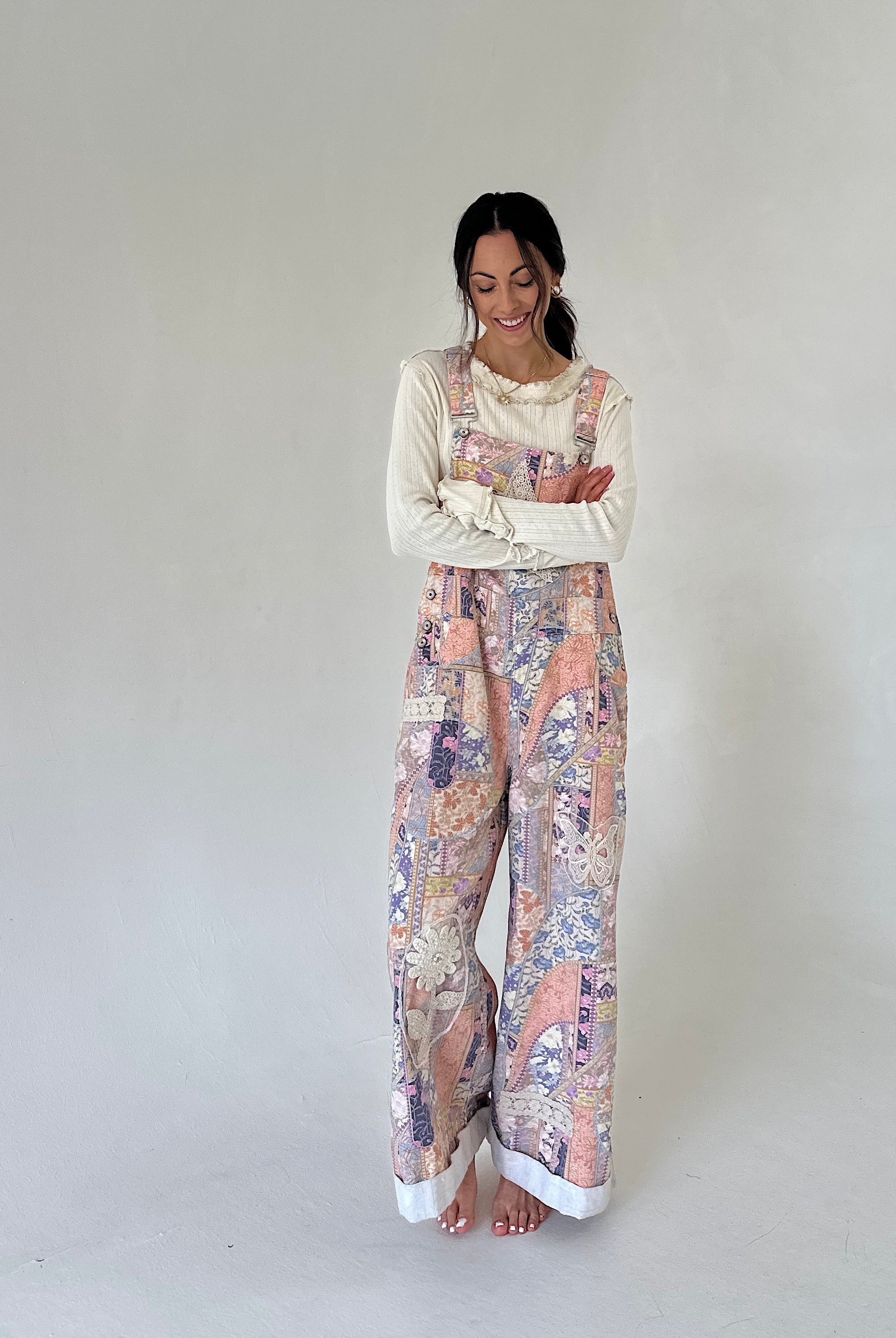 A woman stands barefoot against a plain light gray studio background, wearing a cream long-sleeve top layered under pastel multicolor wide-leg overalls with a floral and abstract print.