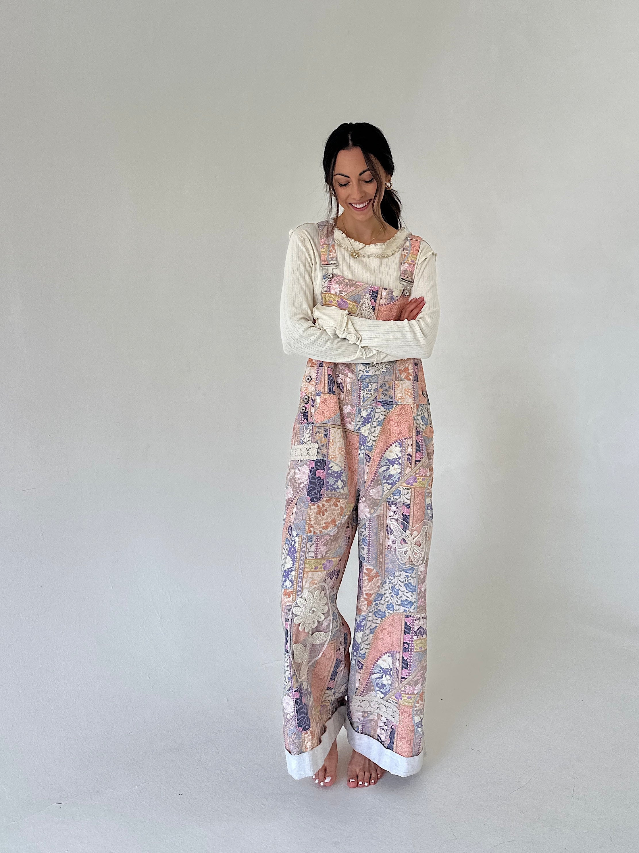 A woman stands barefoot against a plain light gray studio background, wearing a cream long-sleeve top layered under pastel multicolor wide-leg overalls with a floral and abstract print.