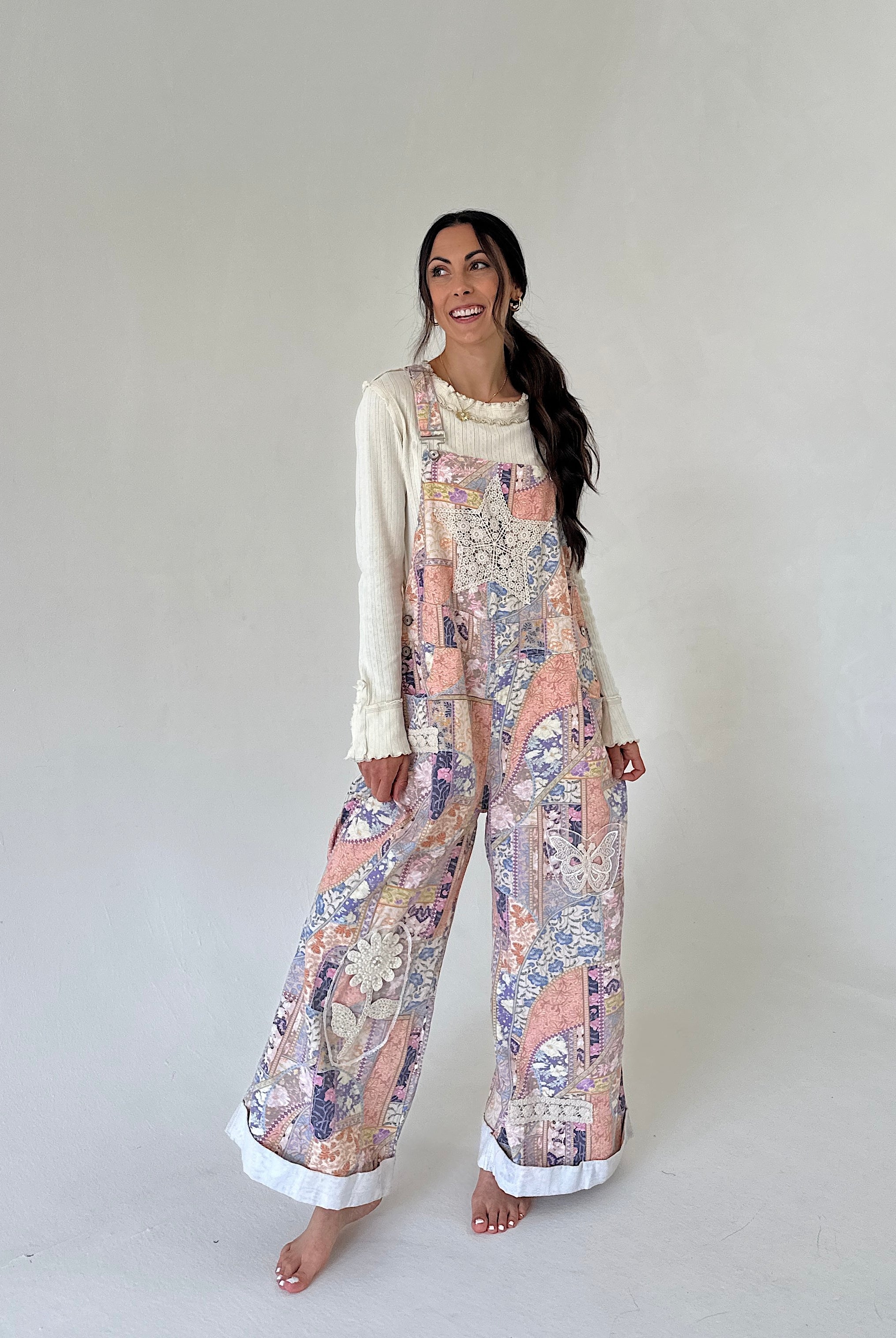 A woman modeling a cream long-sleeve top worn under pastel multicolor patchwork-print overall pants with white cuffed hems, standing against a plain light gray studio background.