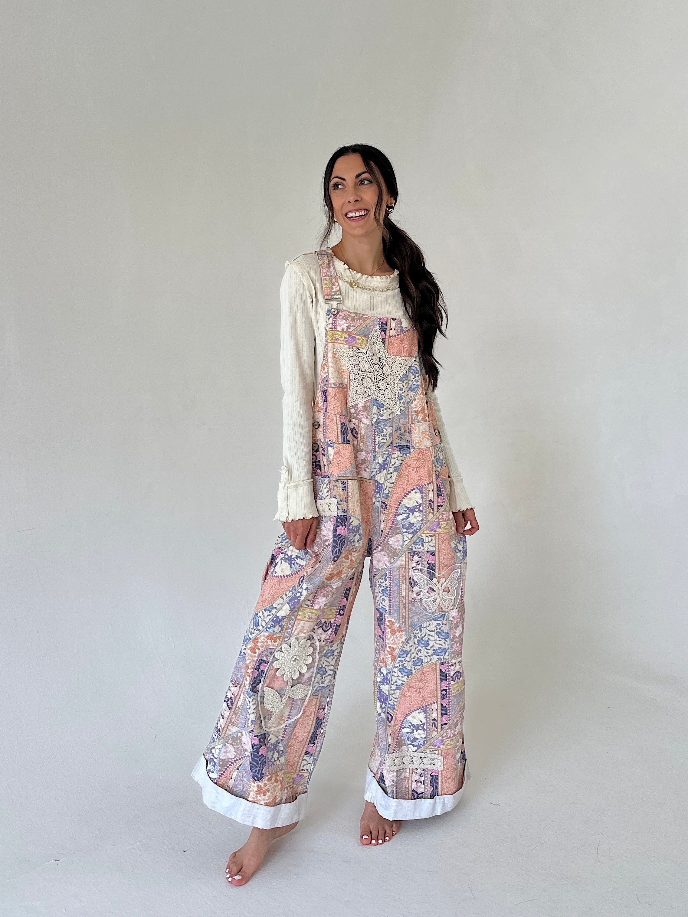 A woman modeling a cream long-sleeve top worn under pastel multicolor patchwork-print overall pants with white cuffed hems, standing against a plain light gray studio background.