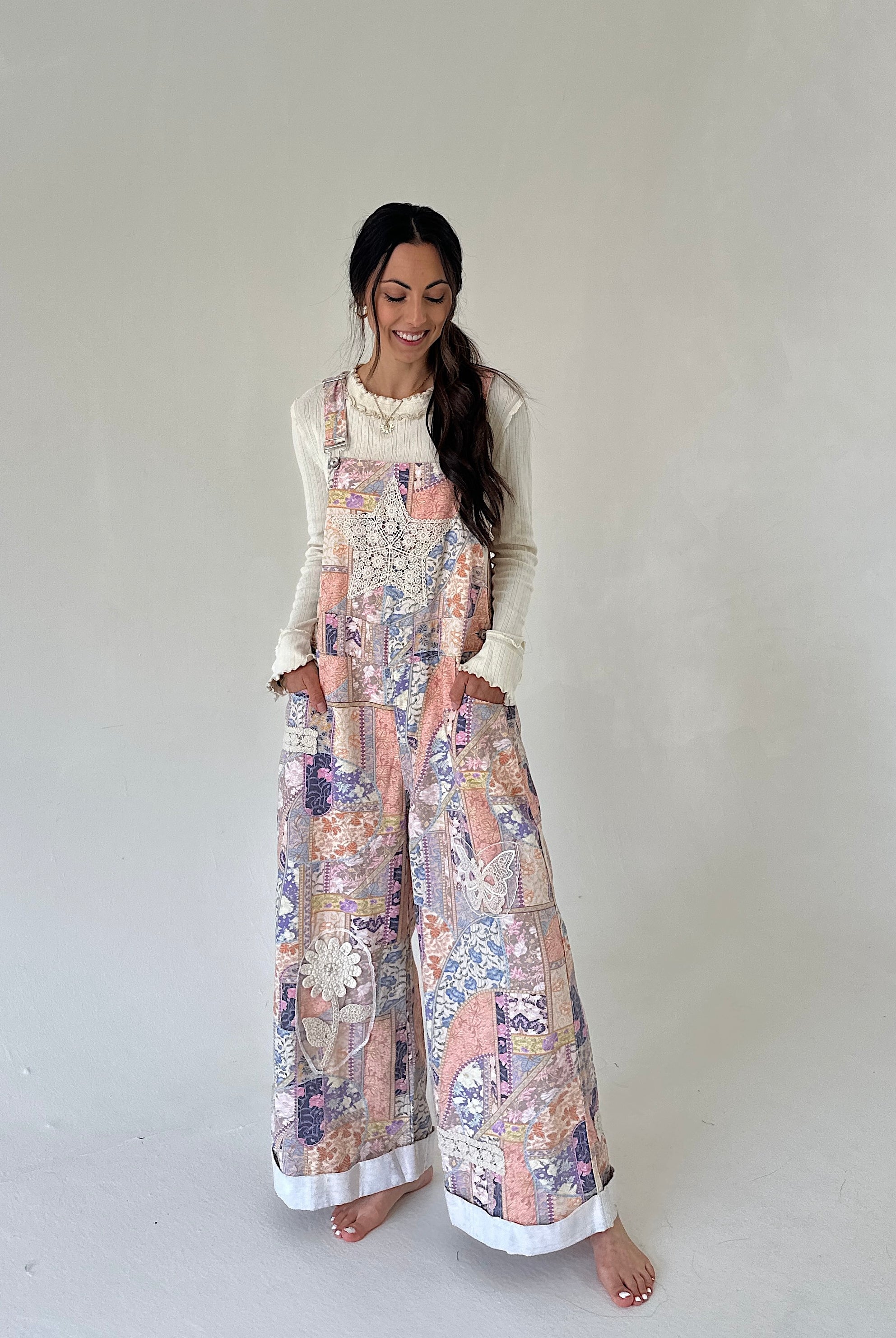 A woman modeling a sleeveless floral print overall jumpsuit with wide legs, worn over a cream long-sleeve top, standing against a plain light gray studio background.