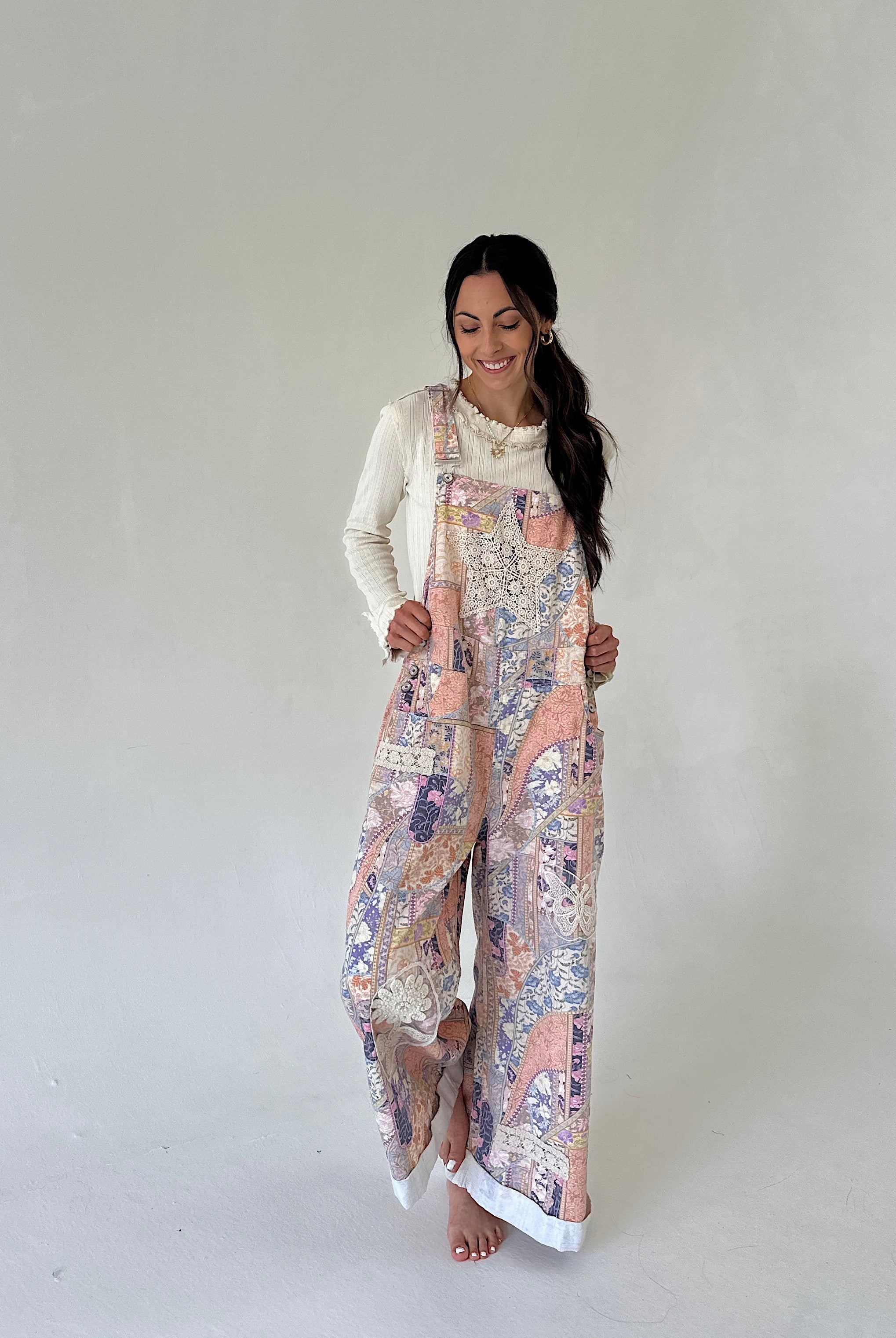 A woman modeling a pair of wide-leg jumpsuit overalls with a multicolor paisley and floral print in pink, beige, and purple, worn over a white long-sleeve top against a neutral studio background.