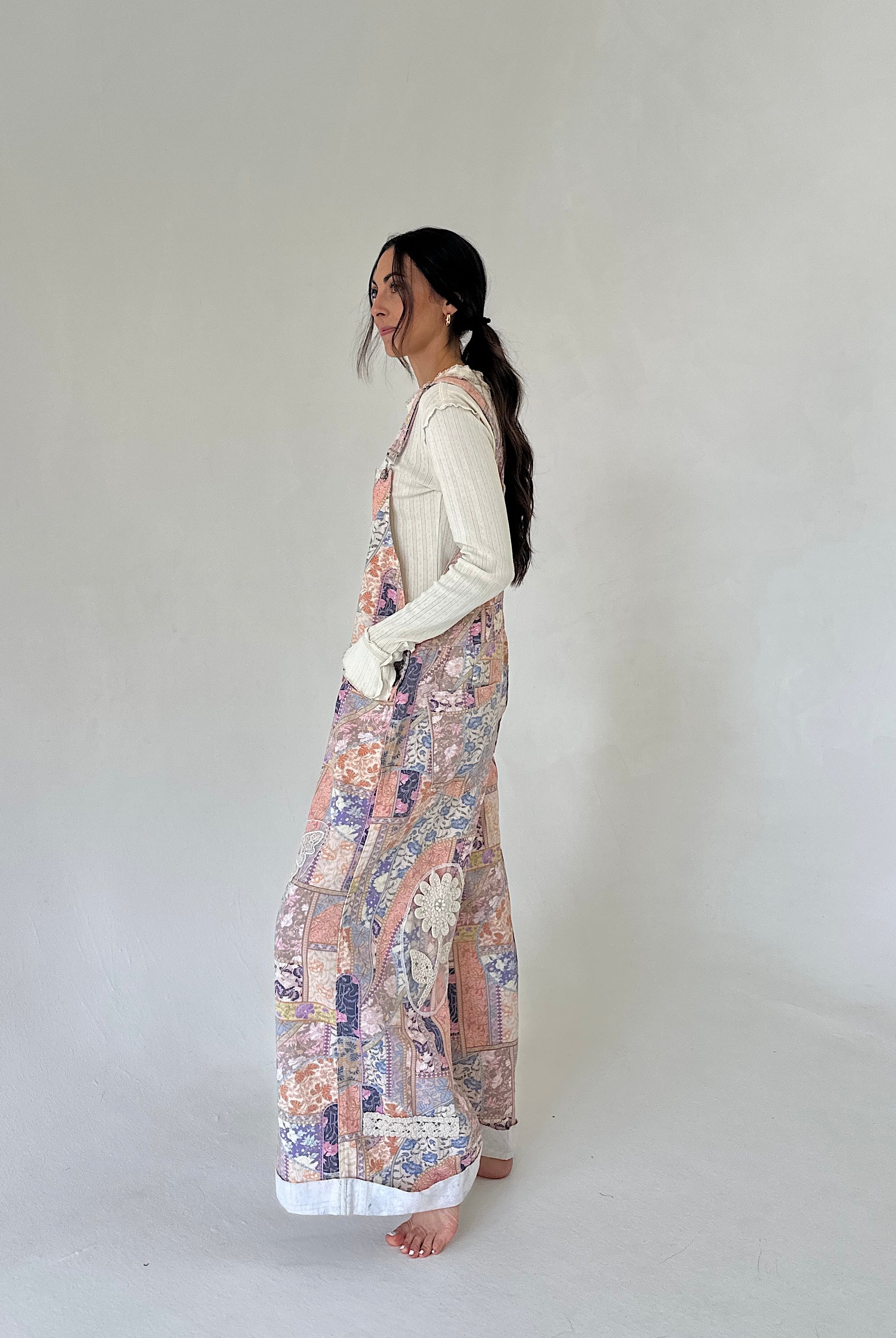 A woman stands in profile wearing a cream ribbed long-sleeve top and wide-leg overalls with a multicolor floral and paisley print in soft pink, purple, blue, and beige tones. She is barefoot against a plain, light gray studio background.