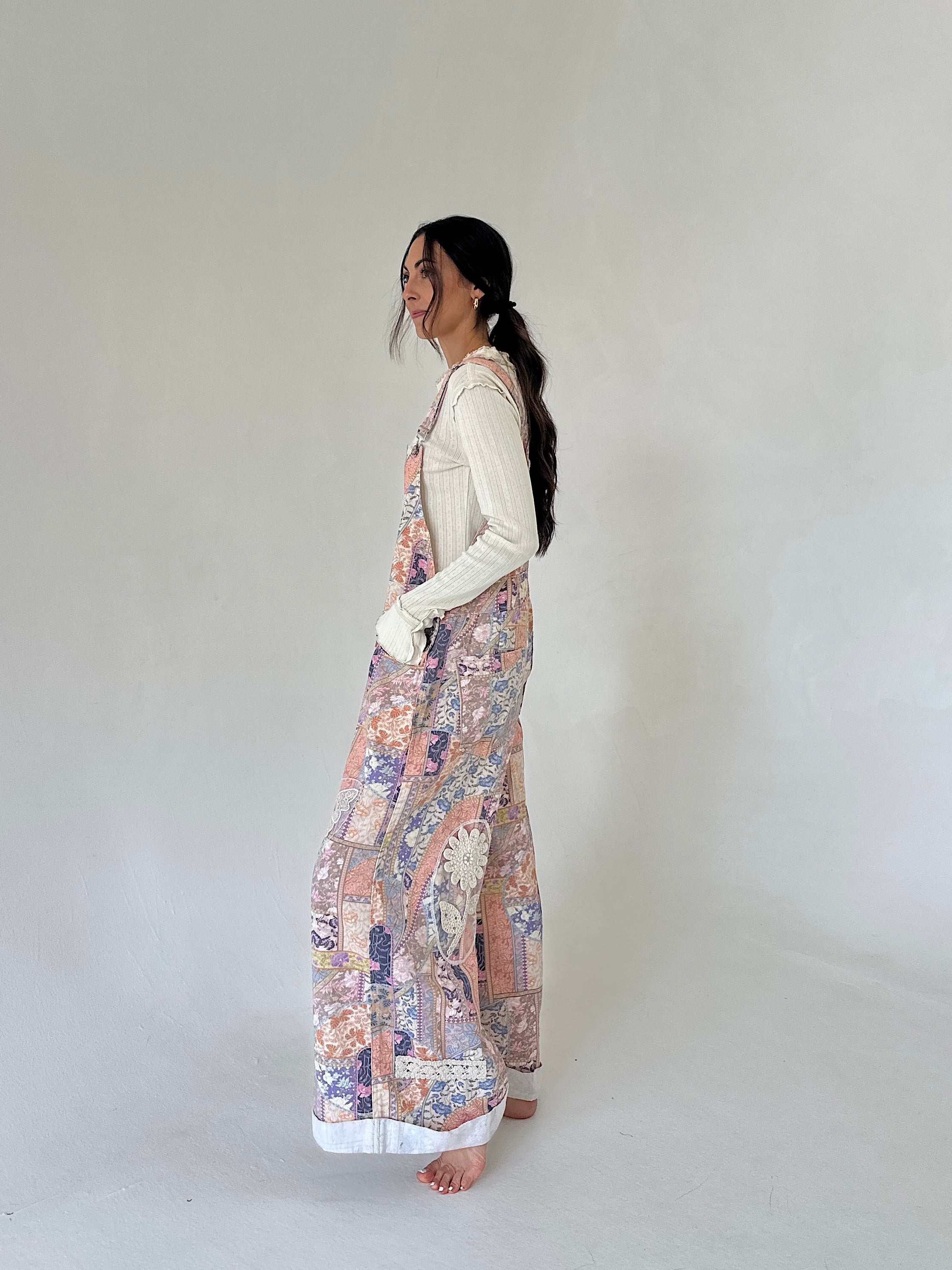 A woman stands in profile wearing a cream ribbed long-sleeve top and wide-leg overalls with a multicolor floral and paisley print in soft pink, purple, blue, and beige tones. She is barefoot against a plain, light gray studio background.
