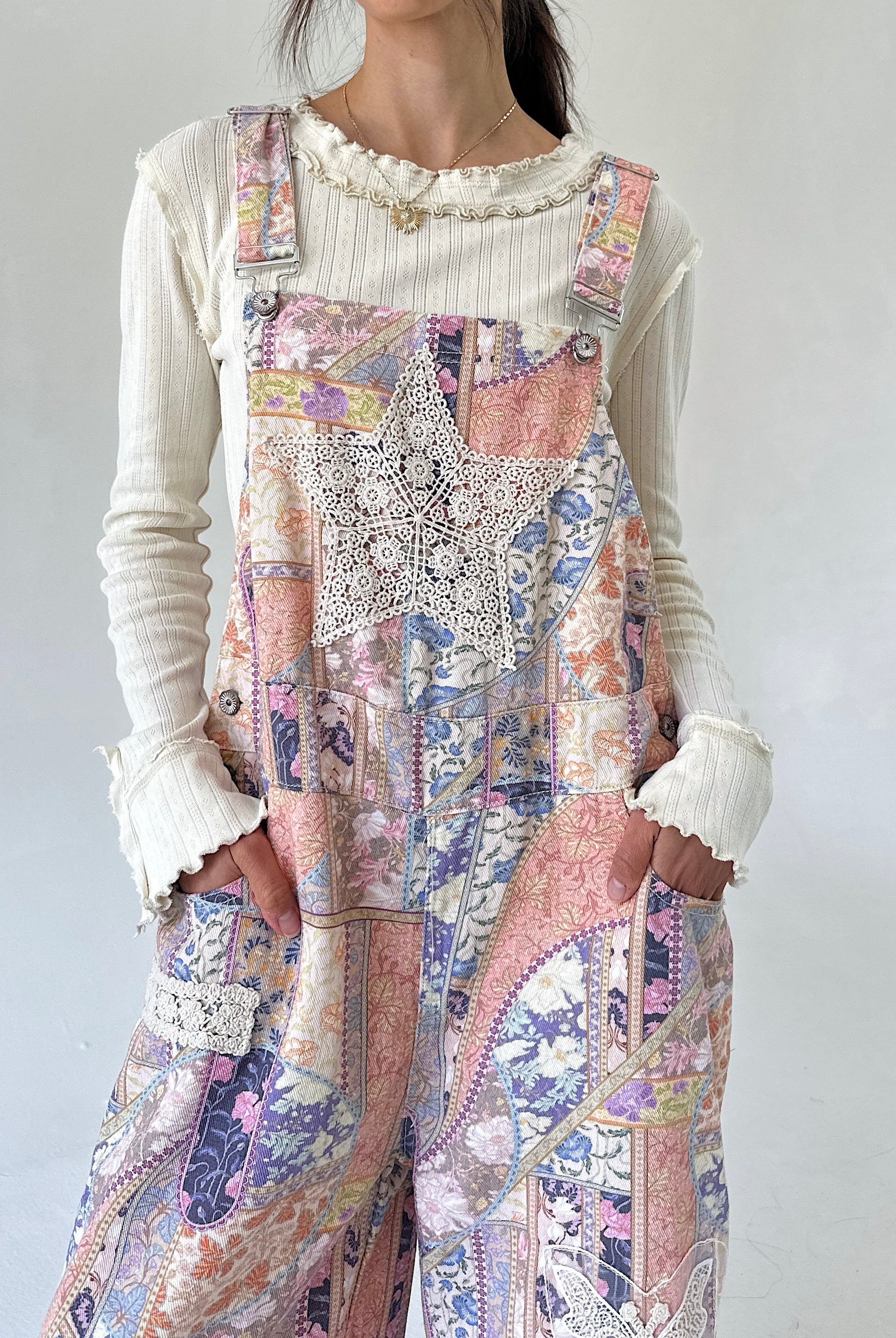 A person wearing a cream ribbed long-sleeve top under pastel patchwork overall pants with lace applique and floral prints in pink, purple, beige, and blue.
