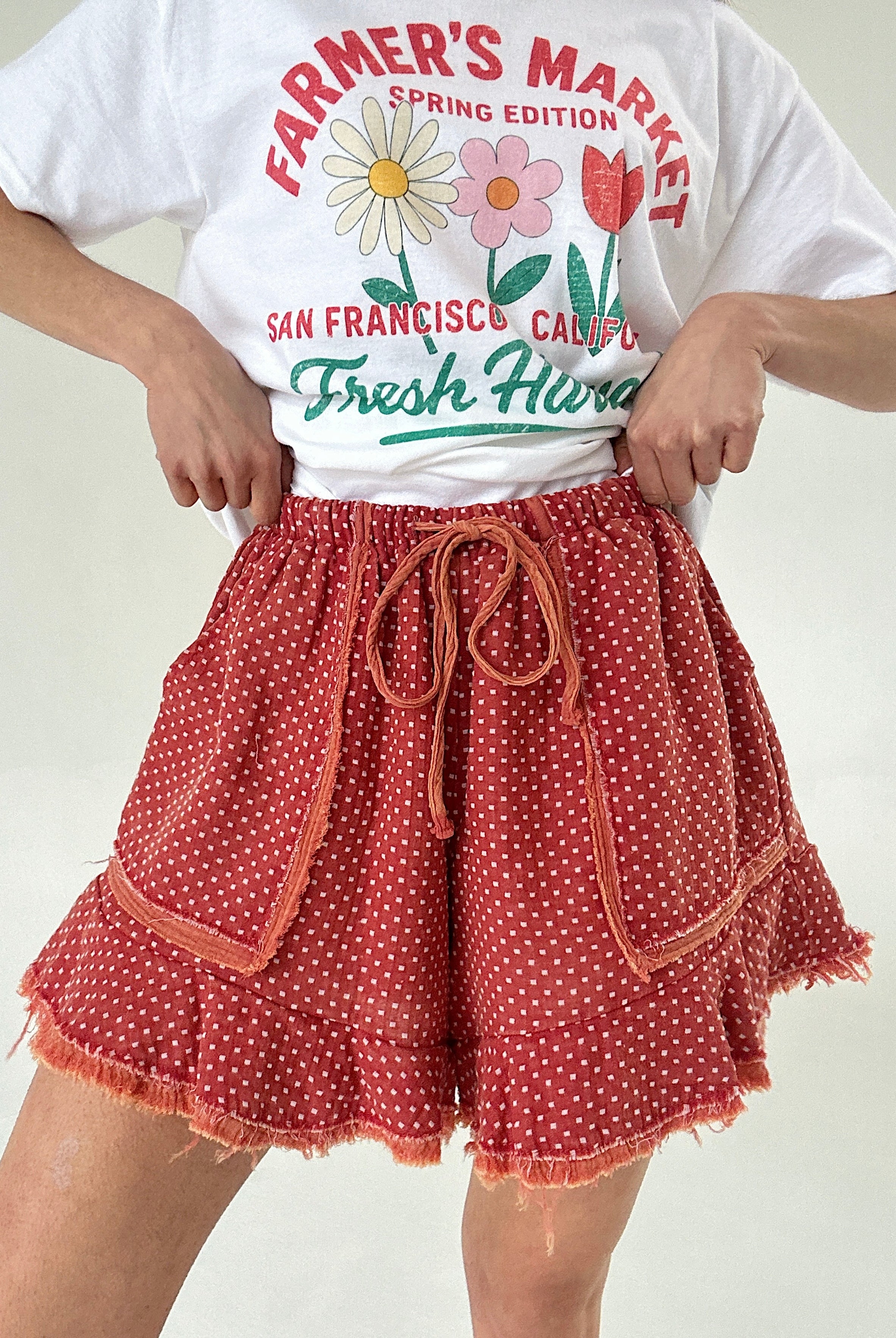 Person wearing a white t-shirt with floral graphics and text, paired with red polka dot shorts on a plain background.