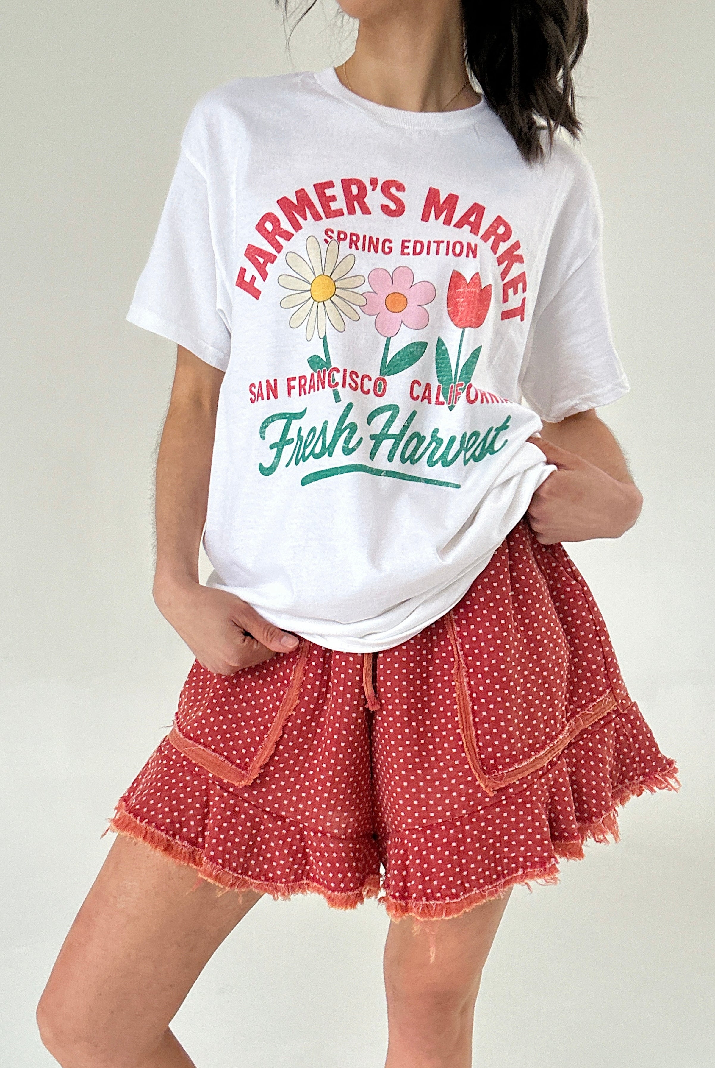 Person wearing a white t-shirt with 'Farmer's Market' text and graphics, paired with red polka dot shorts.