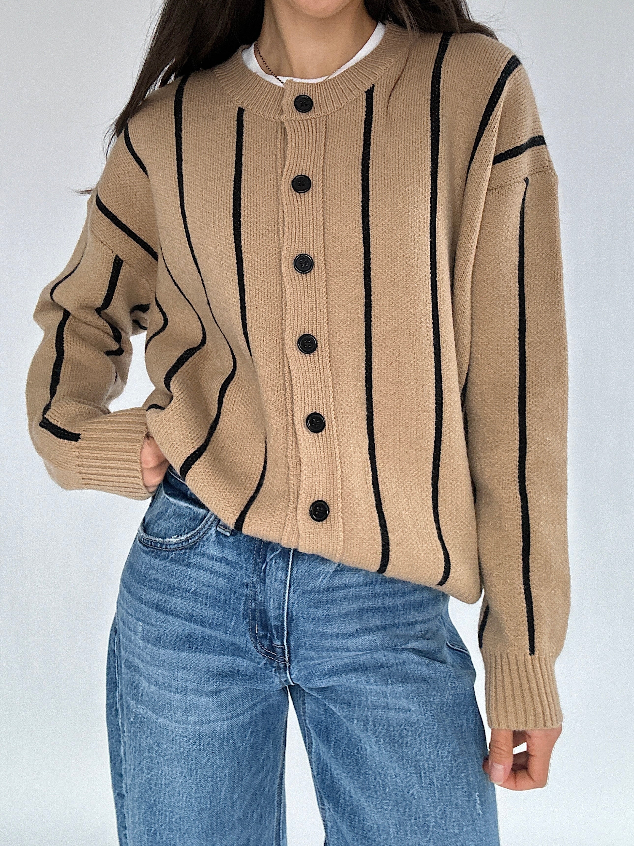 Person wearing a beige sweater with black stripes and buttons, paired with blue jeans on a white background
